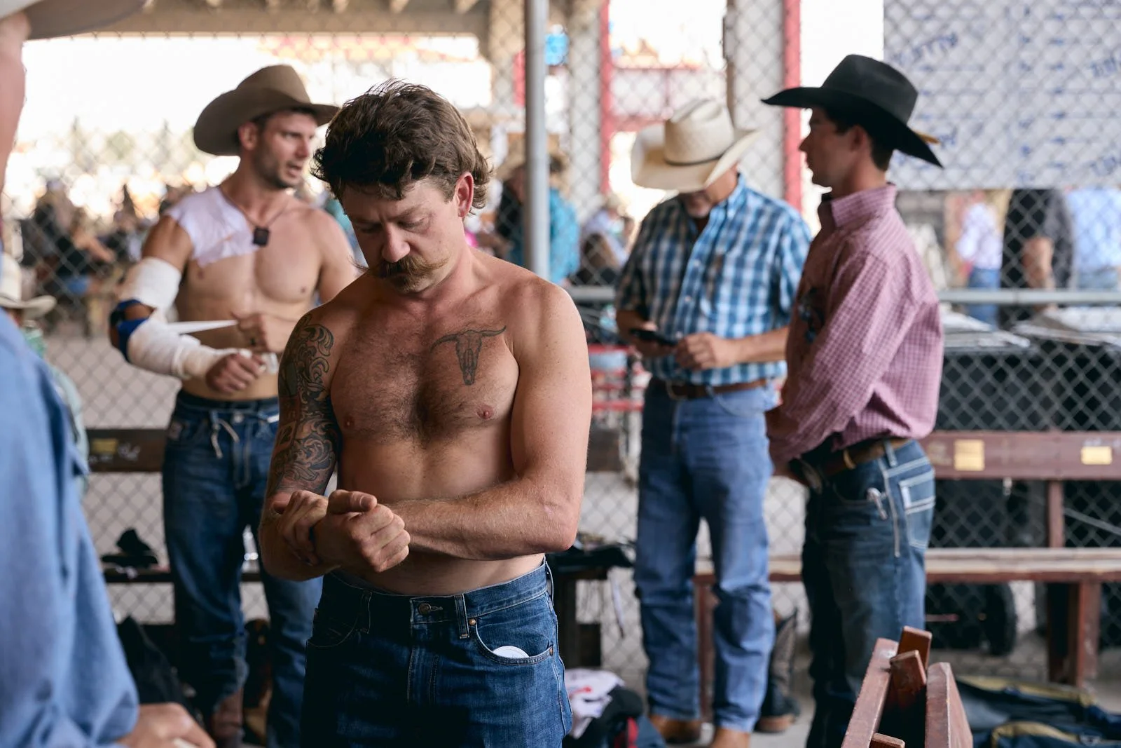 A shirtless man with tattoos on his chest and arm standing outdoors at a rodeo staging area, adjusting or inspecting his arm.