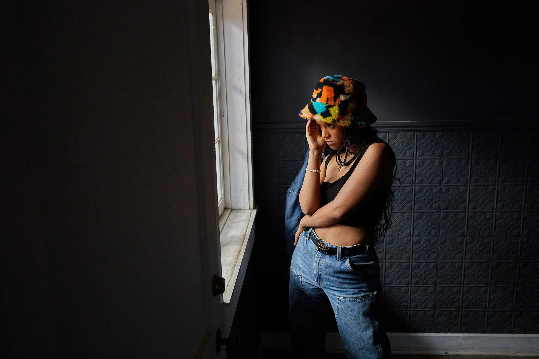 Model with a colorful hat posing in a dark room looking out a window