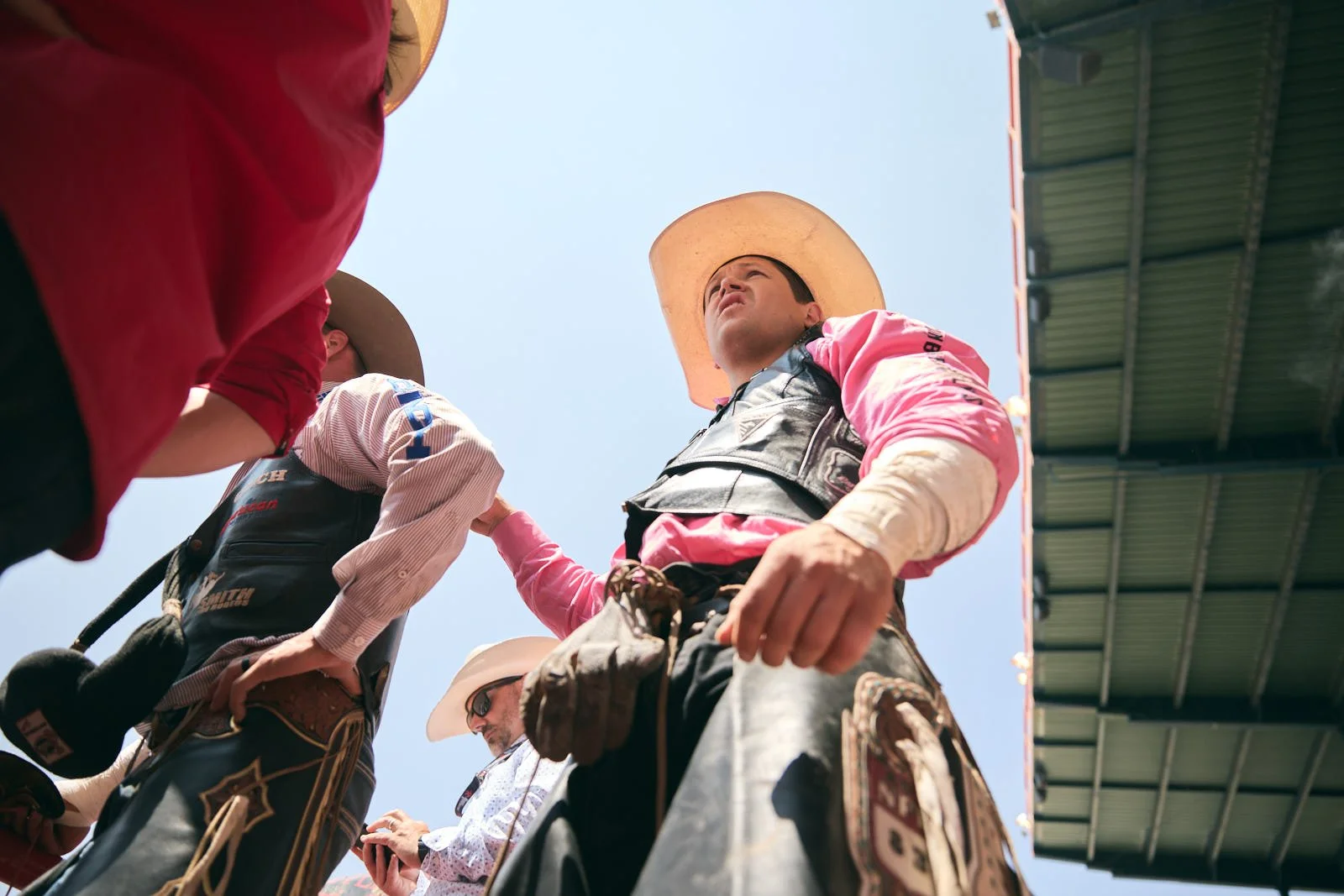 Group of people dressed in cowboy attire, including hats and western wear, gathered outdoors with a clear sky overhead, a rodeo