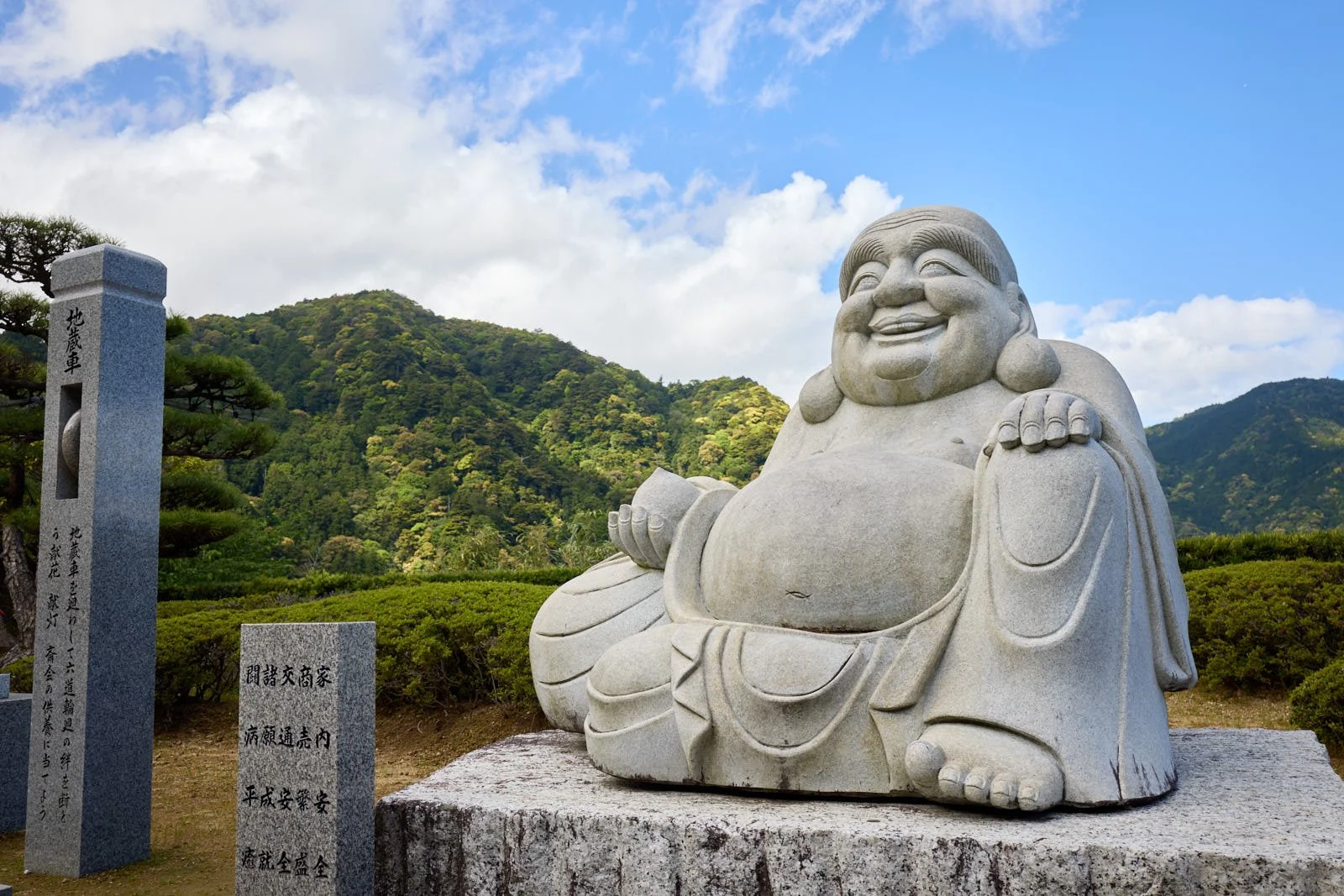 Stone statue of a laughing, plump, Buddha with earrings, sitting with one knee up, outside against a backdrop of green mountains and a partly cloudy sky.