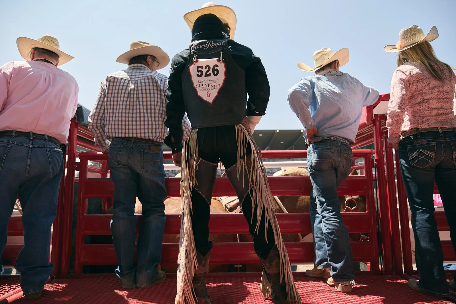 Rodeo assistants and rider wearing cowboy hats and jeans standing behind red chute at rodeo under clear blue sky.