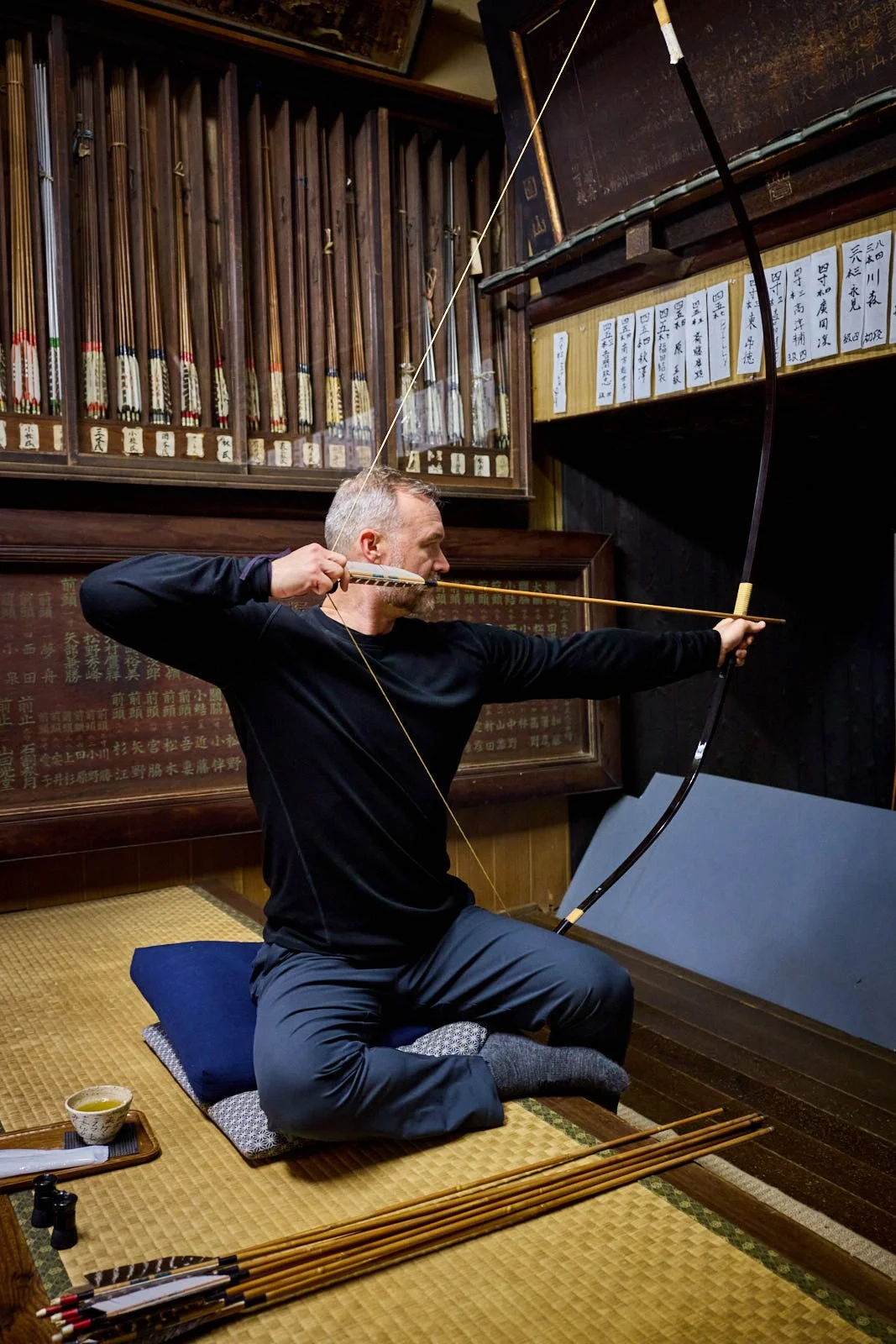 Man dressed in black practicing archery indoors, sitting on a cushion on tatami mat with traditional Japanese weapons and artwork on the walls.