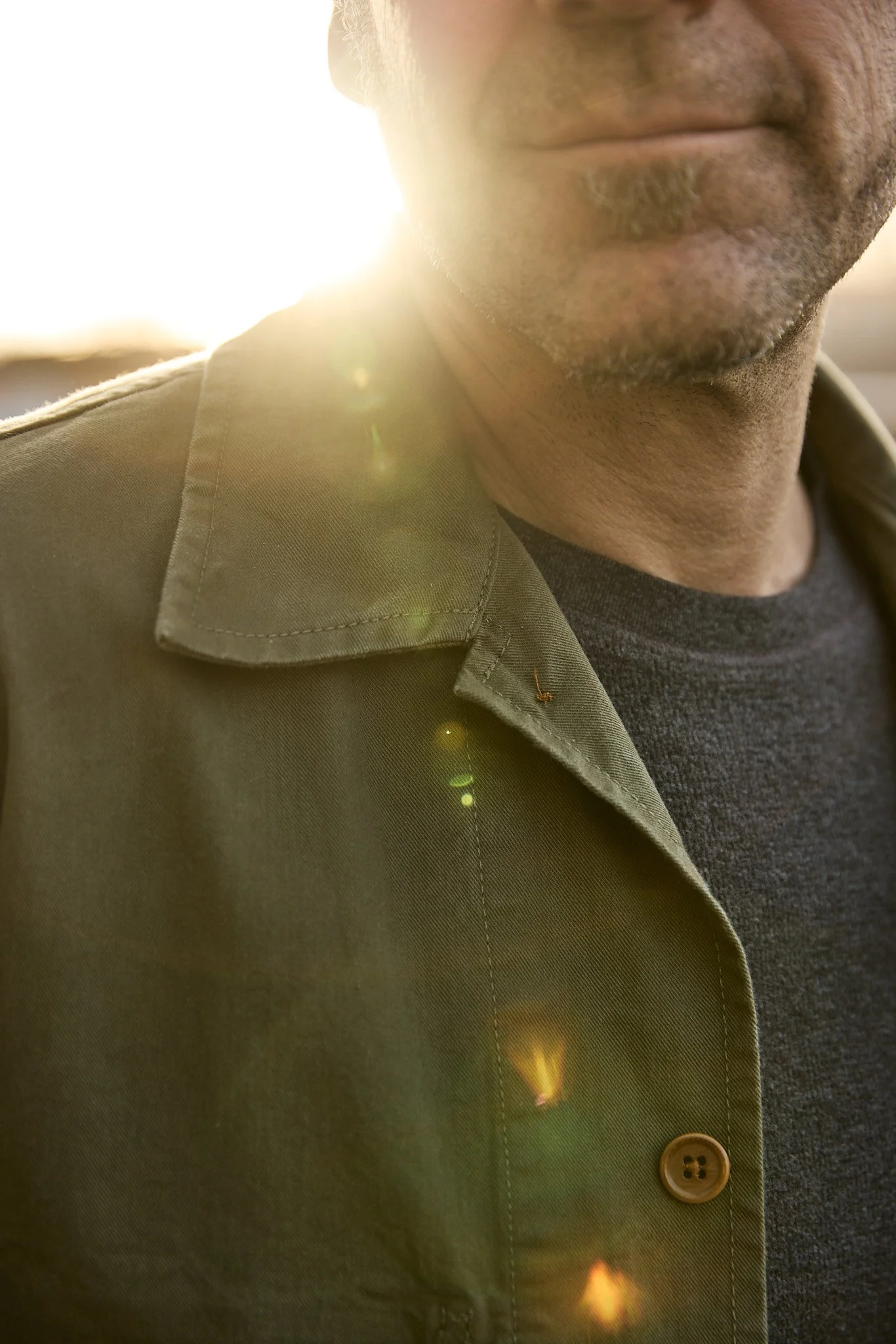 Close-up of a man wearing an olive green jacket and black shirt, with sunlight in the background creating a lens flare effect.