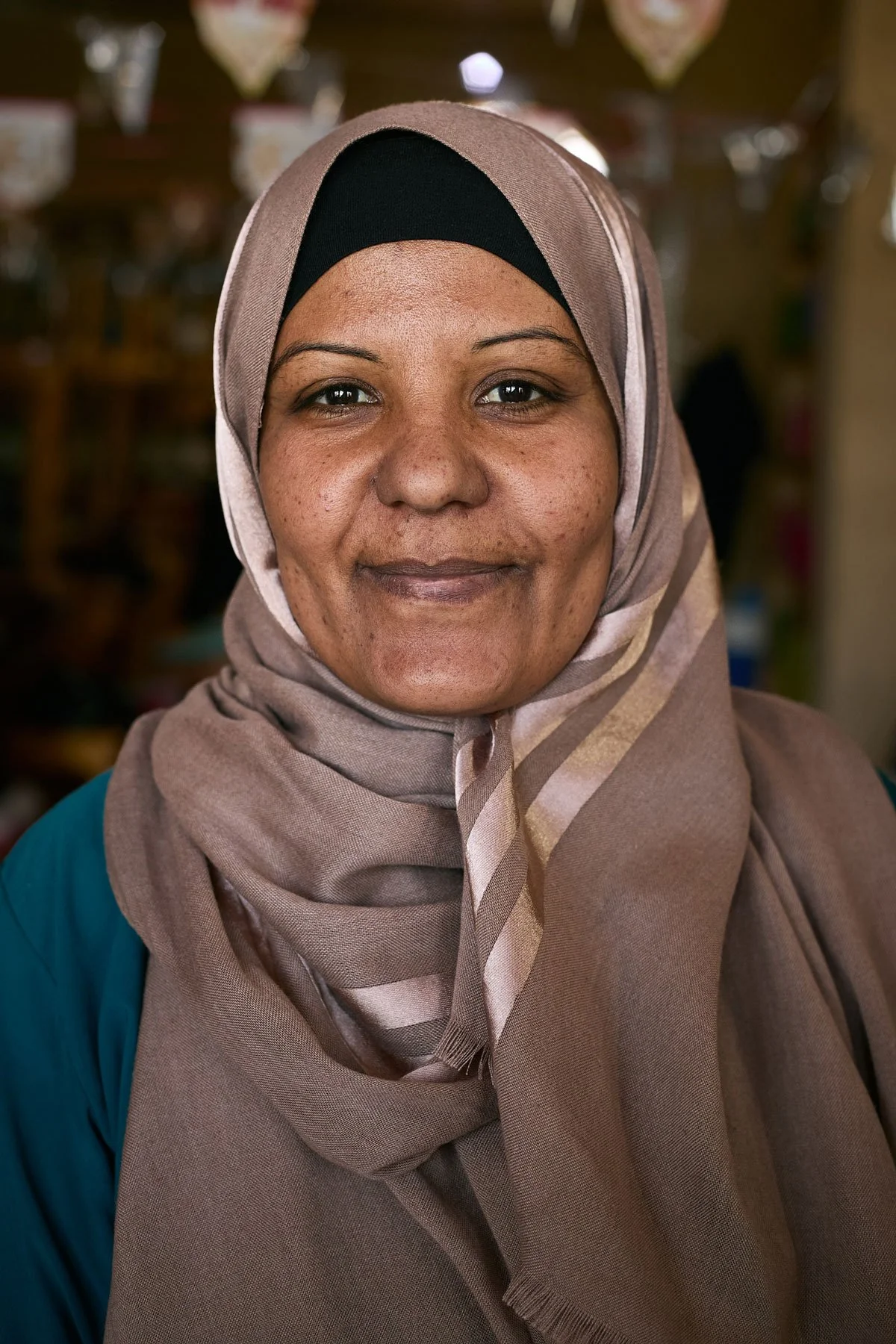 Close-up of a woman wearing a tan hijab with black and beige accents, smiling gently, in a blurred indoor background.