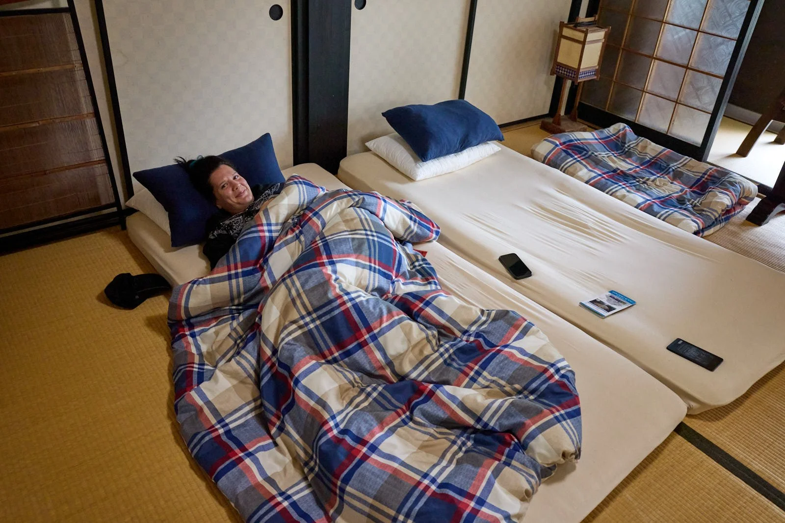 Person lying in bed with stack of cushions, phone, remote control, and magazine on mattress, traditional Japanese room with tatami flooring, sliding doors, and shoji screens.