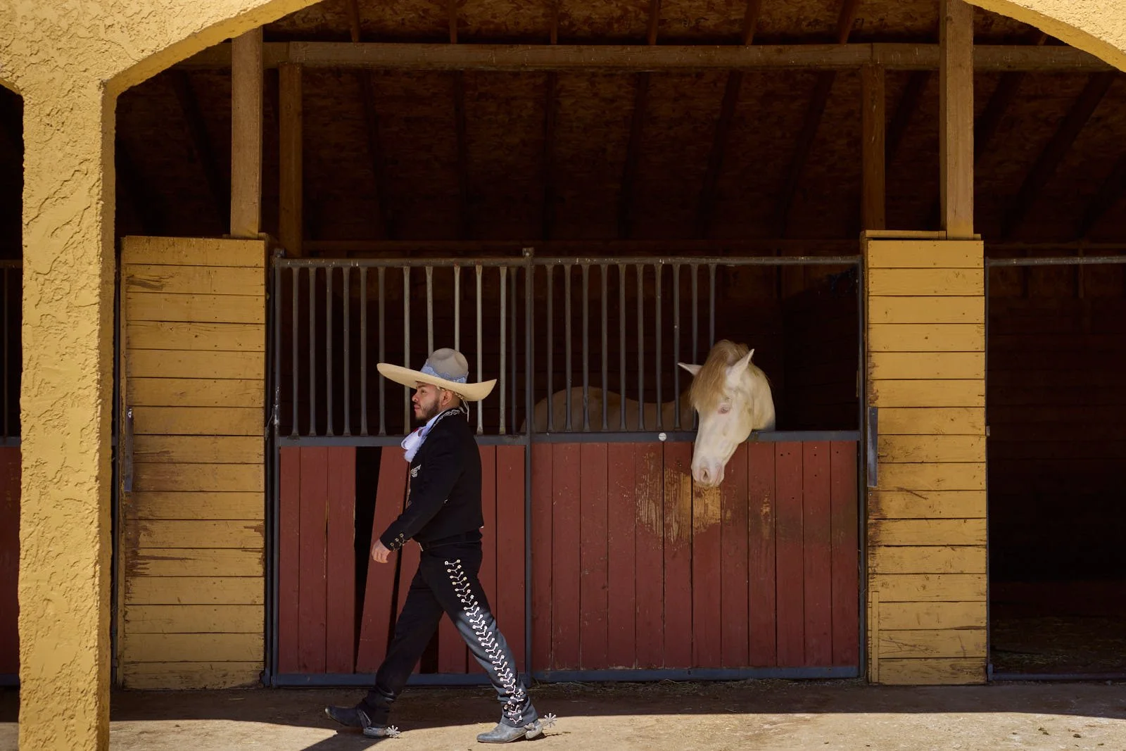 A man dressed in traditional Mexican attire, including a large sombrero and a black suit with silver embroidery, walks past a horse stable with a white horse resting its head on the stable door.