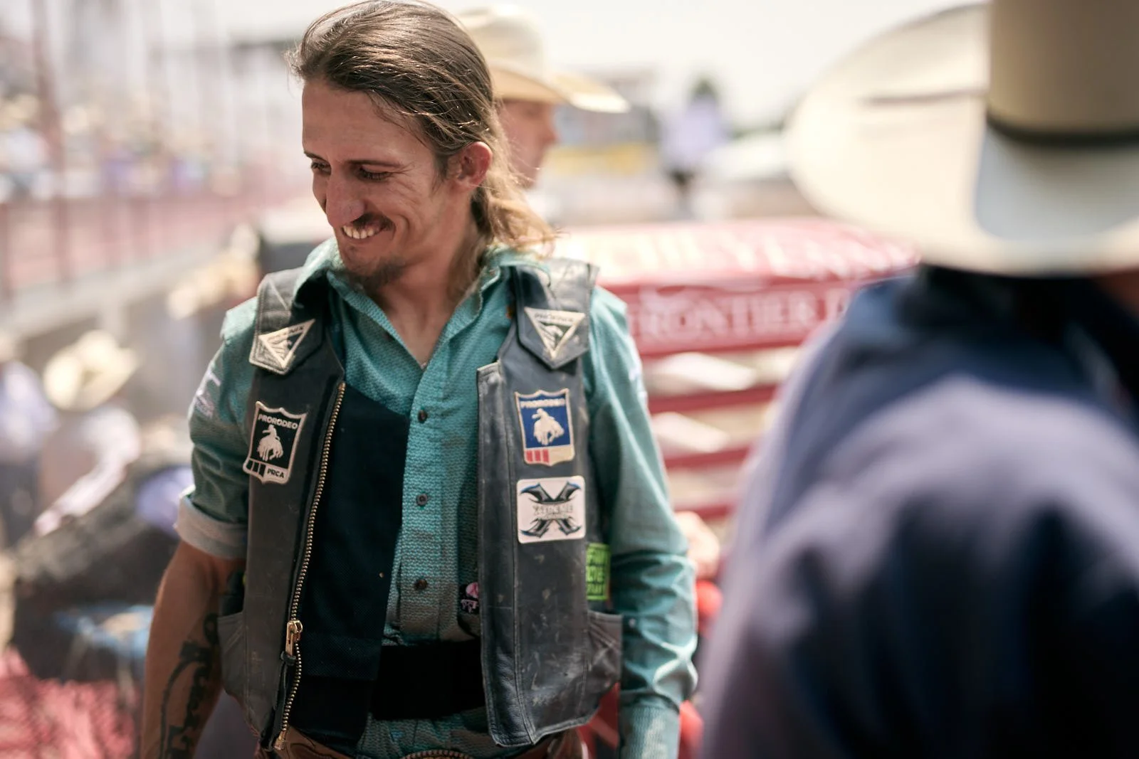 A man with long hair, a beard, and a mustache smiling, wearing a vest with patches, standing outdoors at a market or street fair.