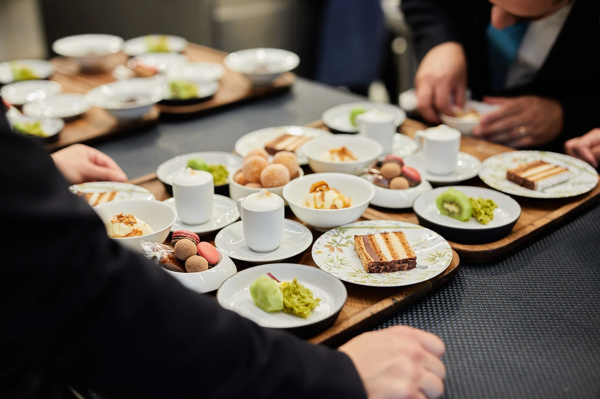 Several plates and bowls with desserts like cake, fruit, and macarons on a wooden table, with people reaching for the treats.