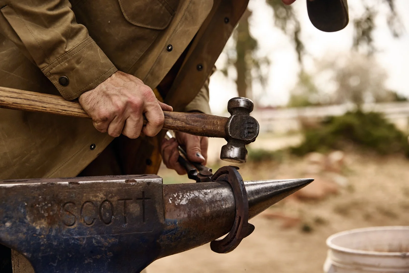A person using a hammer to shape or forge a horseshoe in an outdoor setting, with trees and a bucket in the background.