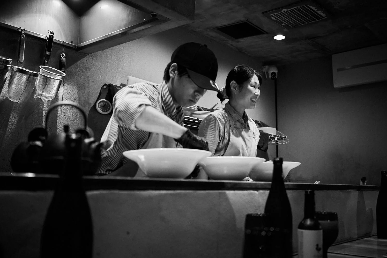 Two people, a man and a woman, working in a restaurant kitchen, preparing food at the counter with bowls in front of them. The scene is in black and white.