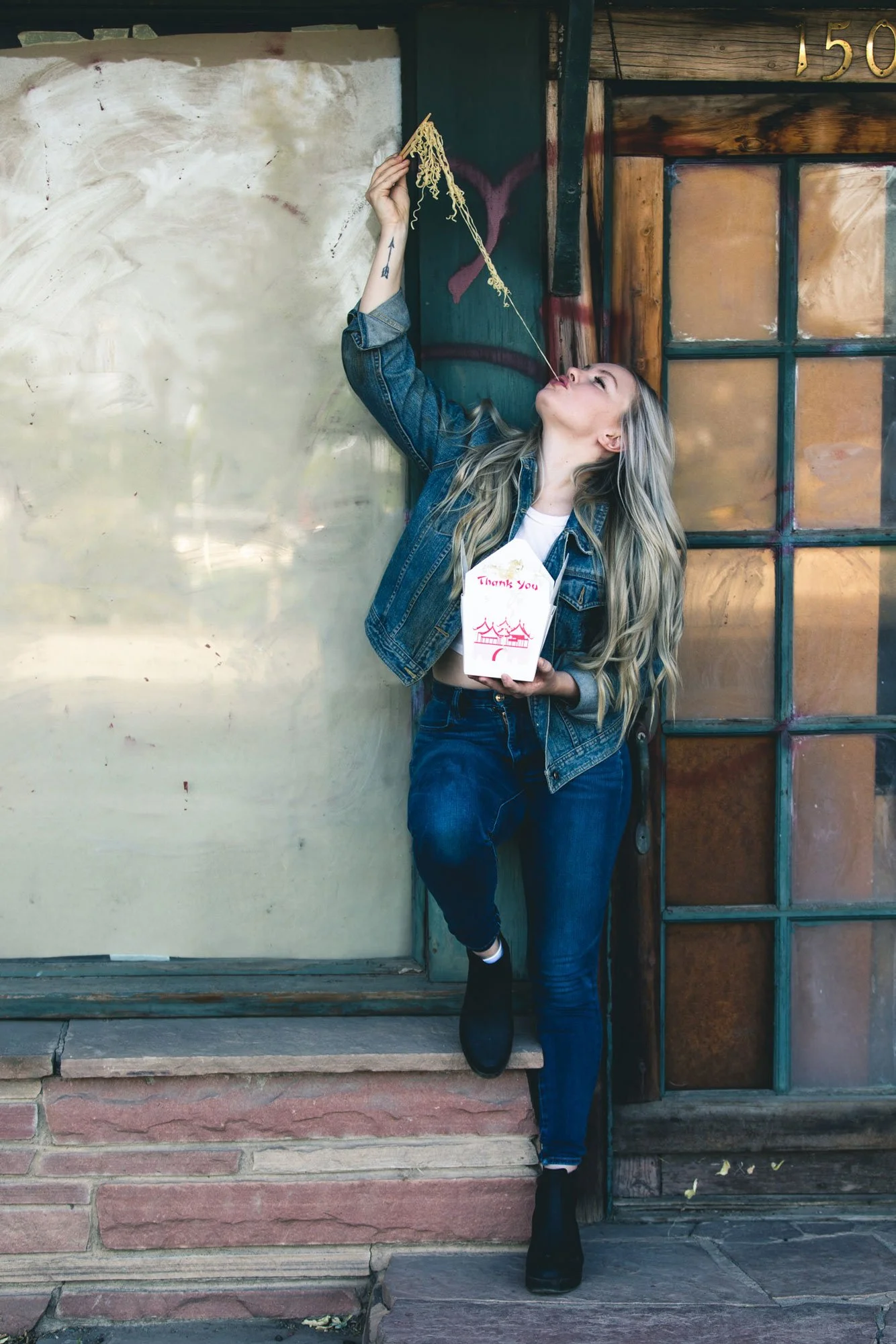 Model standing next to a vacant building eating a long stand of ramen noodles