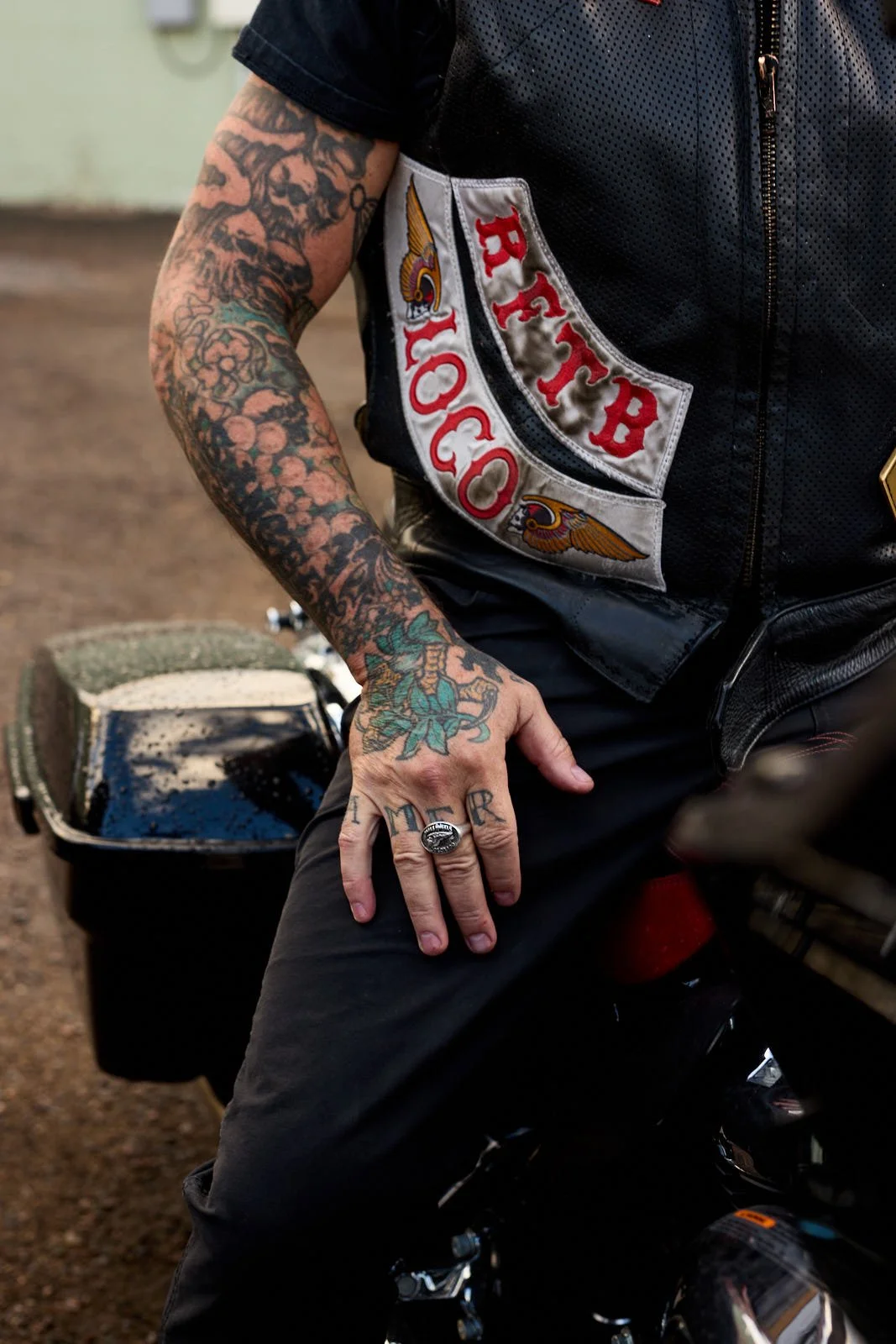 A person with tattooed arms wearing a black vest with patches, resting his hand on this thigh on a motorcycle 