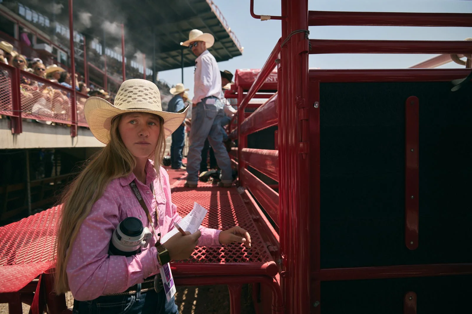 A young girl in a cowboy hat and pink shirt holding a notepad and a camera standing beside a red cattle chute at an outdoor rodeo event with spectators watching from the stands.