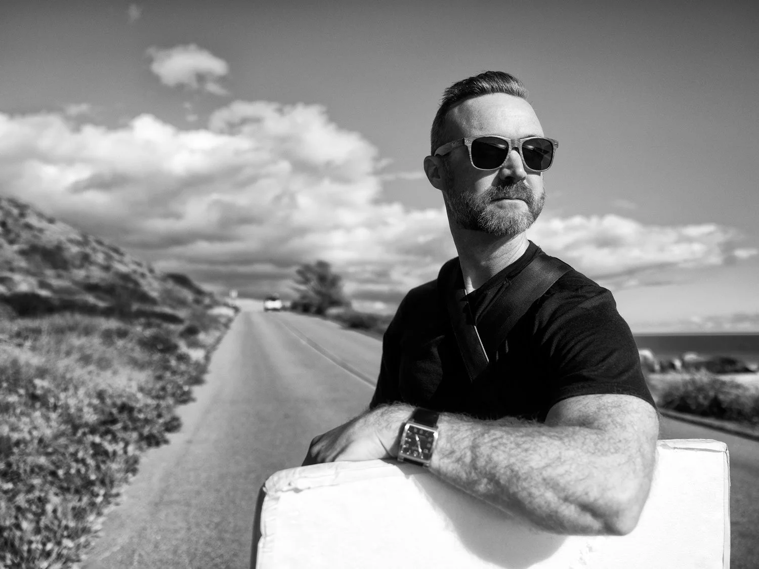 John Robson wearing cheap sunglass sunglasses and a watch resting his arm on a bounce board, standing on a road with a scenic landscape including hills and a cloudy sky in the background.