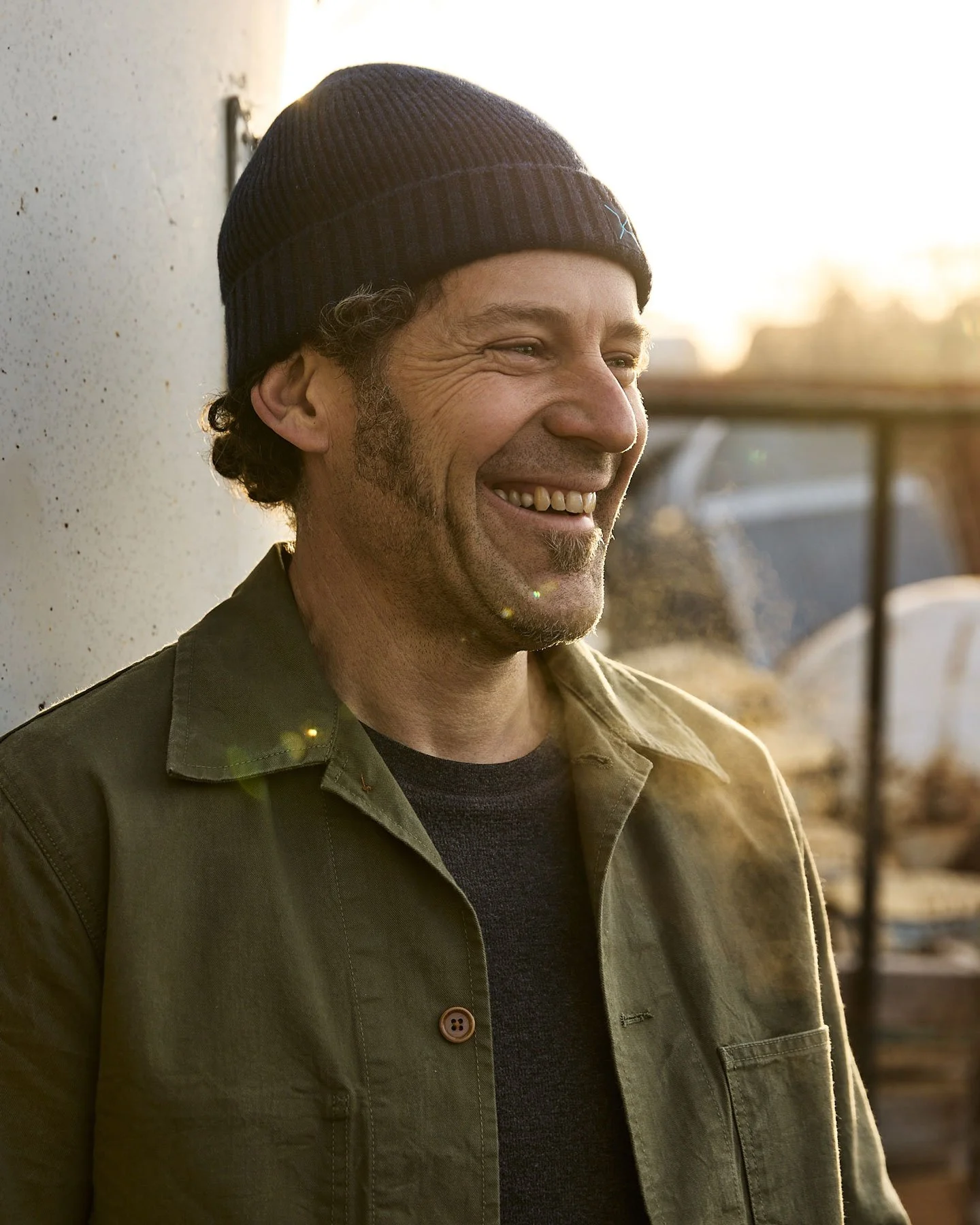 A man with curly hair and facial hair wearing a knit cap, smiling outdoors during sunset.