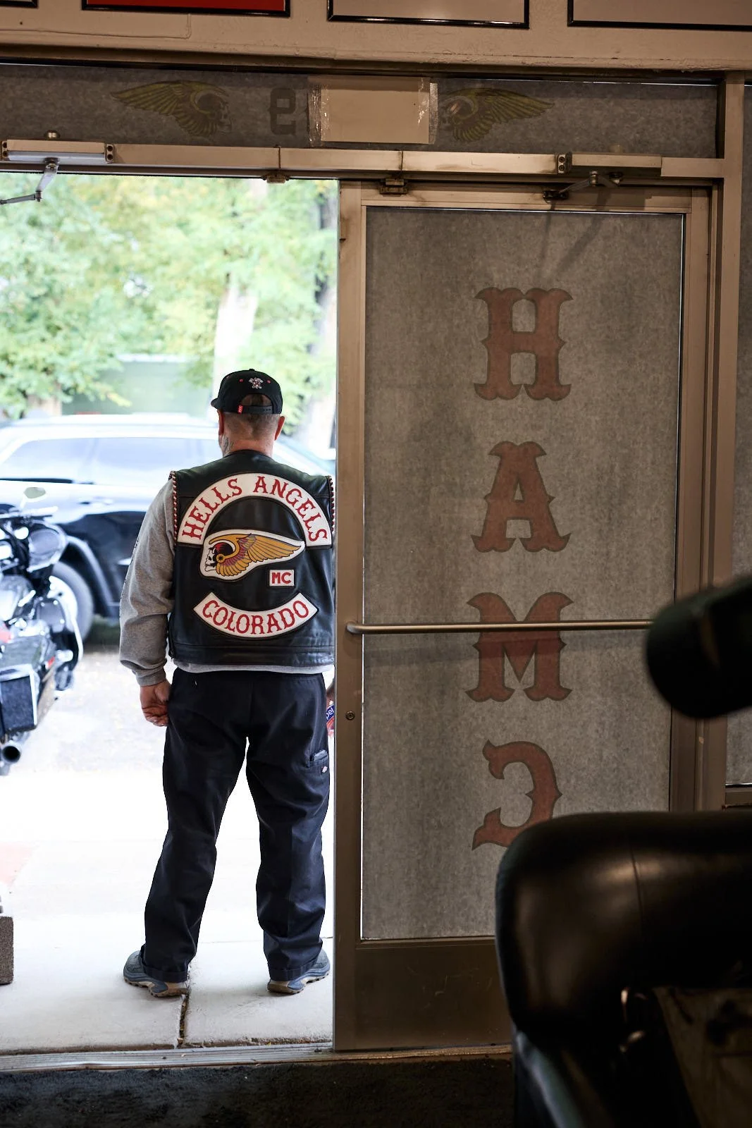 A man dressed in a black vest with a patch that reads 'Hell's Angels Colorado' stands in the doorway of a building, facing outside. Outside, there are trees, a parked black motorcycle, and a black car.