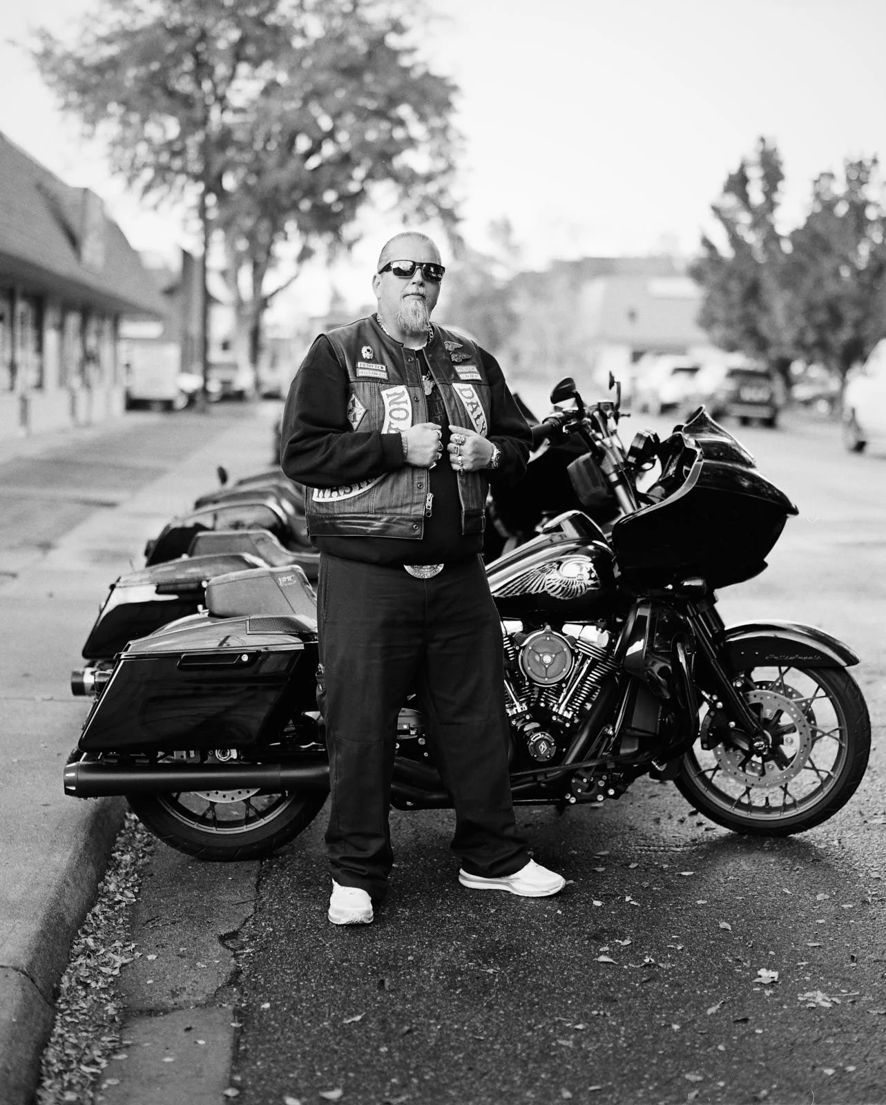 A man wearing sunglasses, a biker vest with patches, and black pants with white shoes stands next to a black motorcycle on a street in a small town.