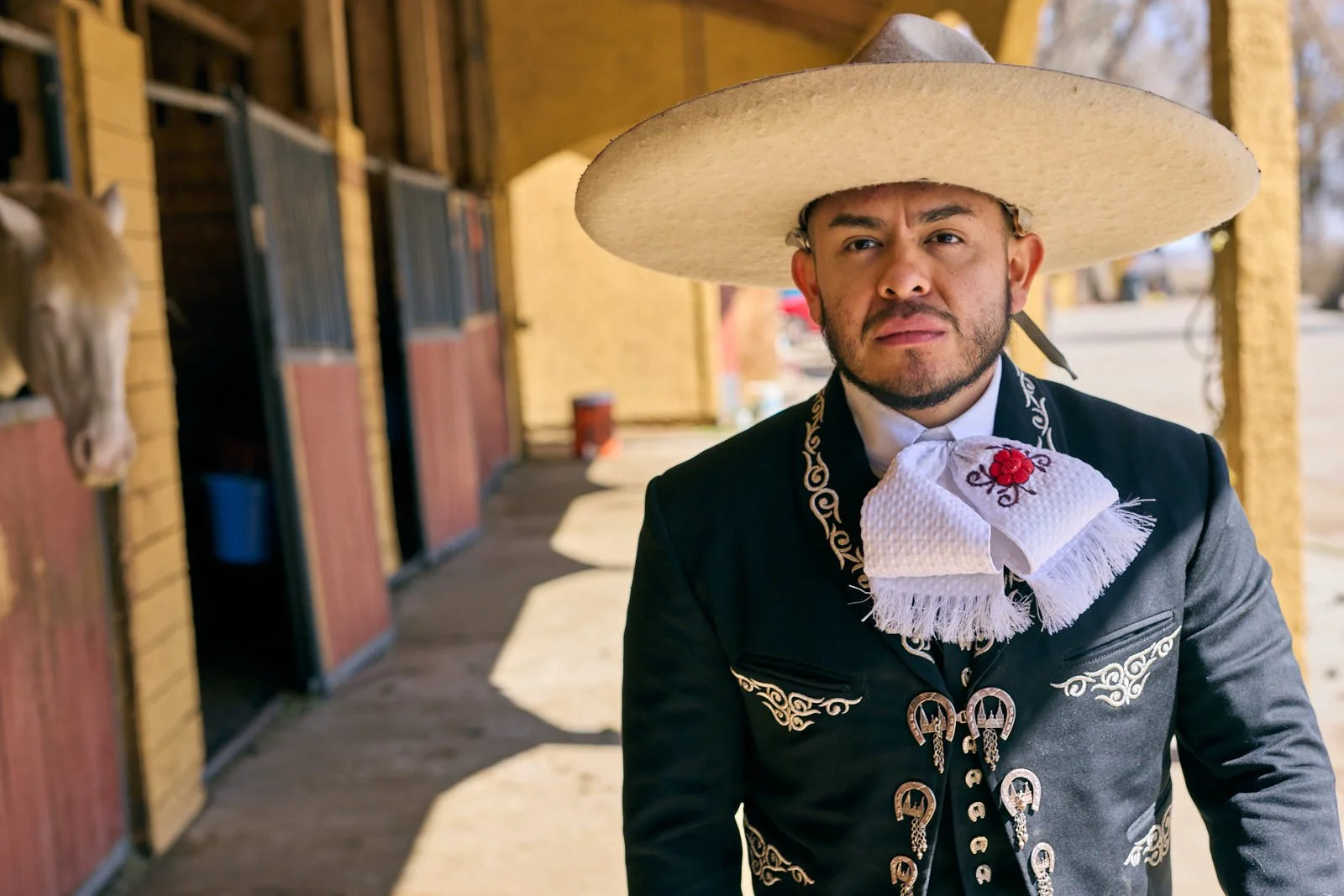A man wearing traditional Mexican charro attire, including a large sombrero and embroidered suit, standing outside a horse stable.