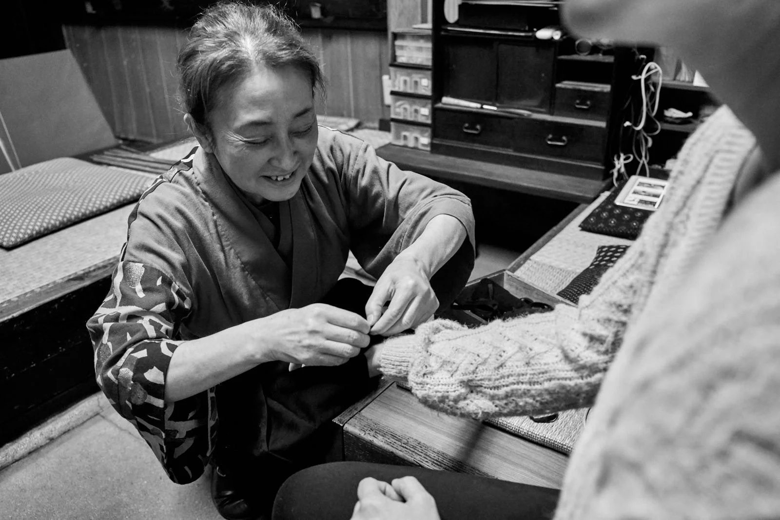 A woman with a big smile, wearing a patterned kimono, is fitting a finger guard on a seated person.