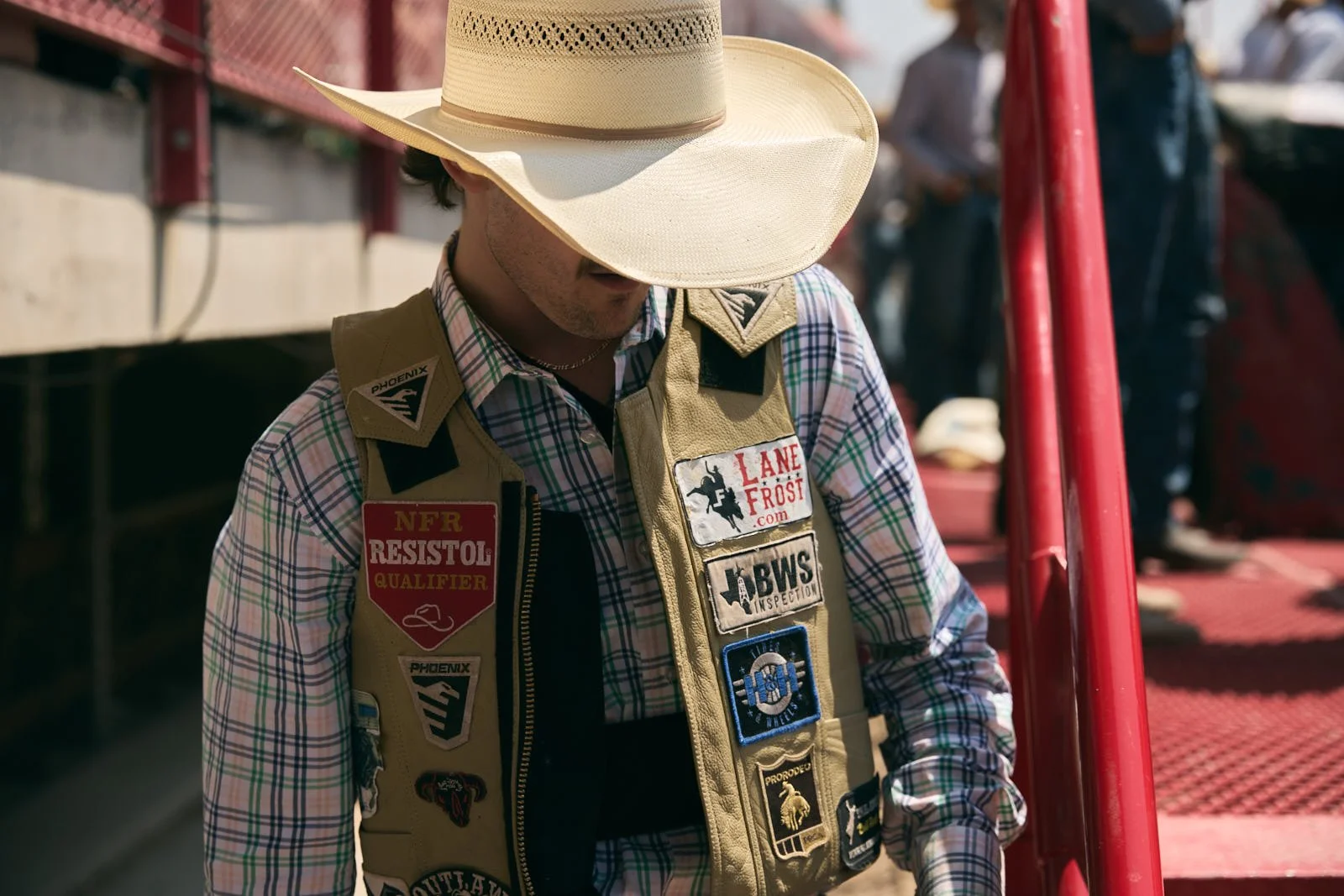 A man wearing a beige vest covered with various patches, a plaid shirt, and a large straw cowboy hat standing near a red metal railing outdoors.