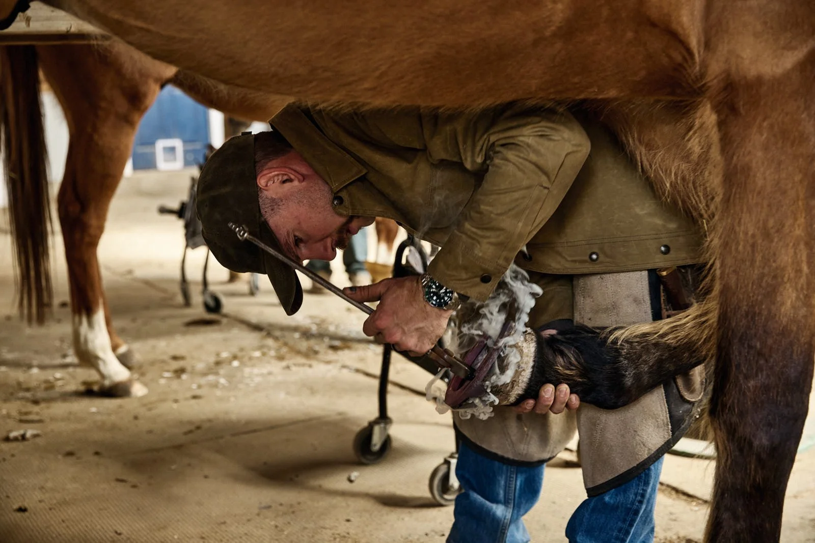 A man hot shoeing a horse's hoof in a barn.