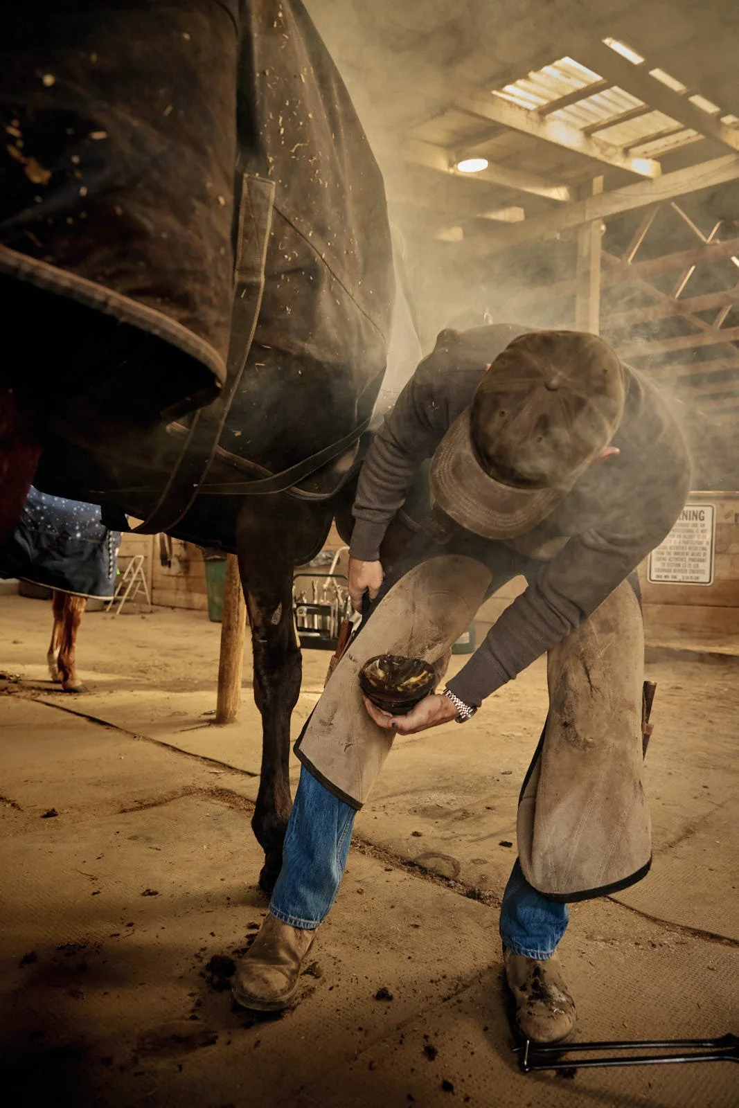 A man wearing a hat and hot shoeing a horse