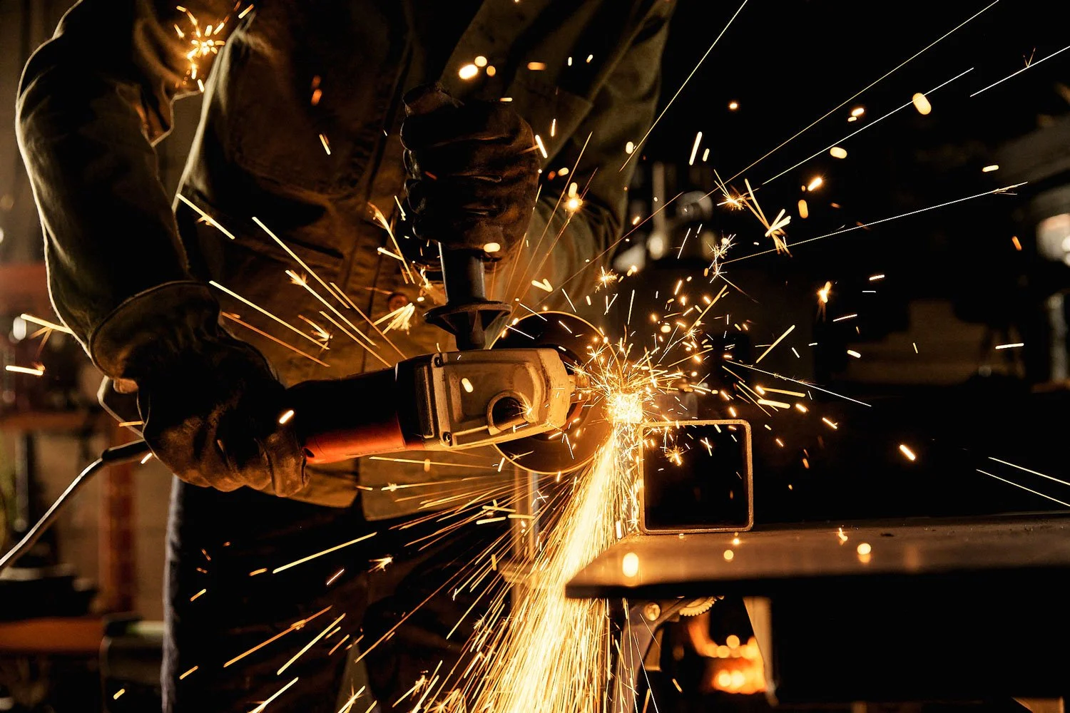A person cutting metal using an angle grinder, sparks flying in multiple directions, in a workshop with tools and equipment in the background.