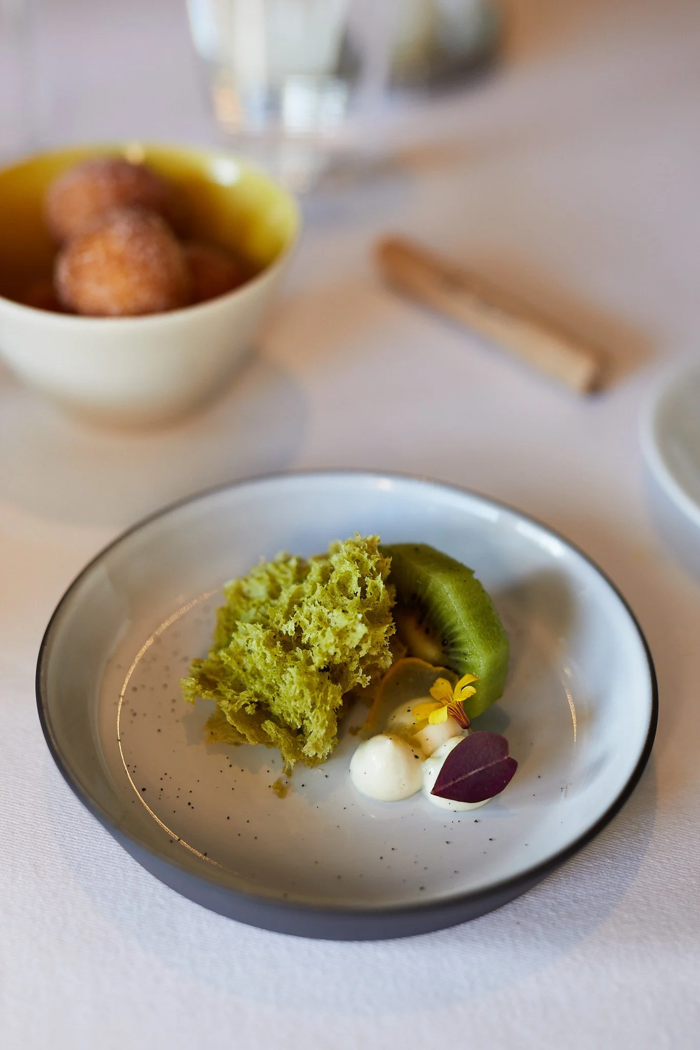 A small dish of green sponge, kiwi slices, dollops of white cream, and edible flowers on a white tablecloth, with a bowl of food and glass of water in the background at the French Laundry.