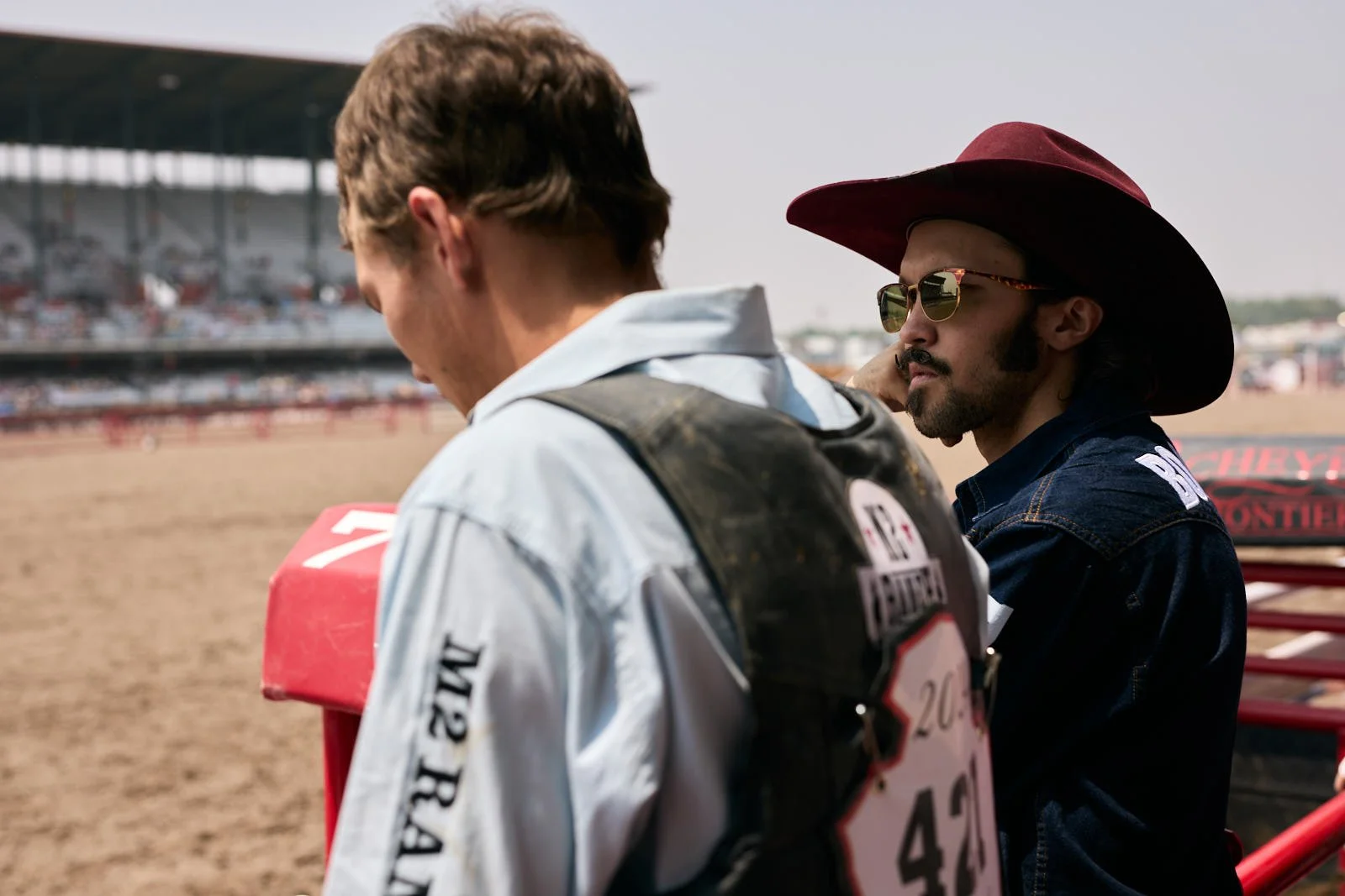 Two men at a rodeo, one wearing a cowboy hat and sunglasses, the other facing away, holding a helmet, with a blurry grandstand in the background.