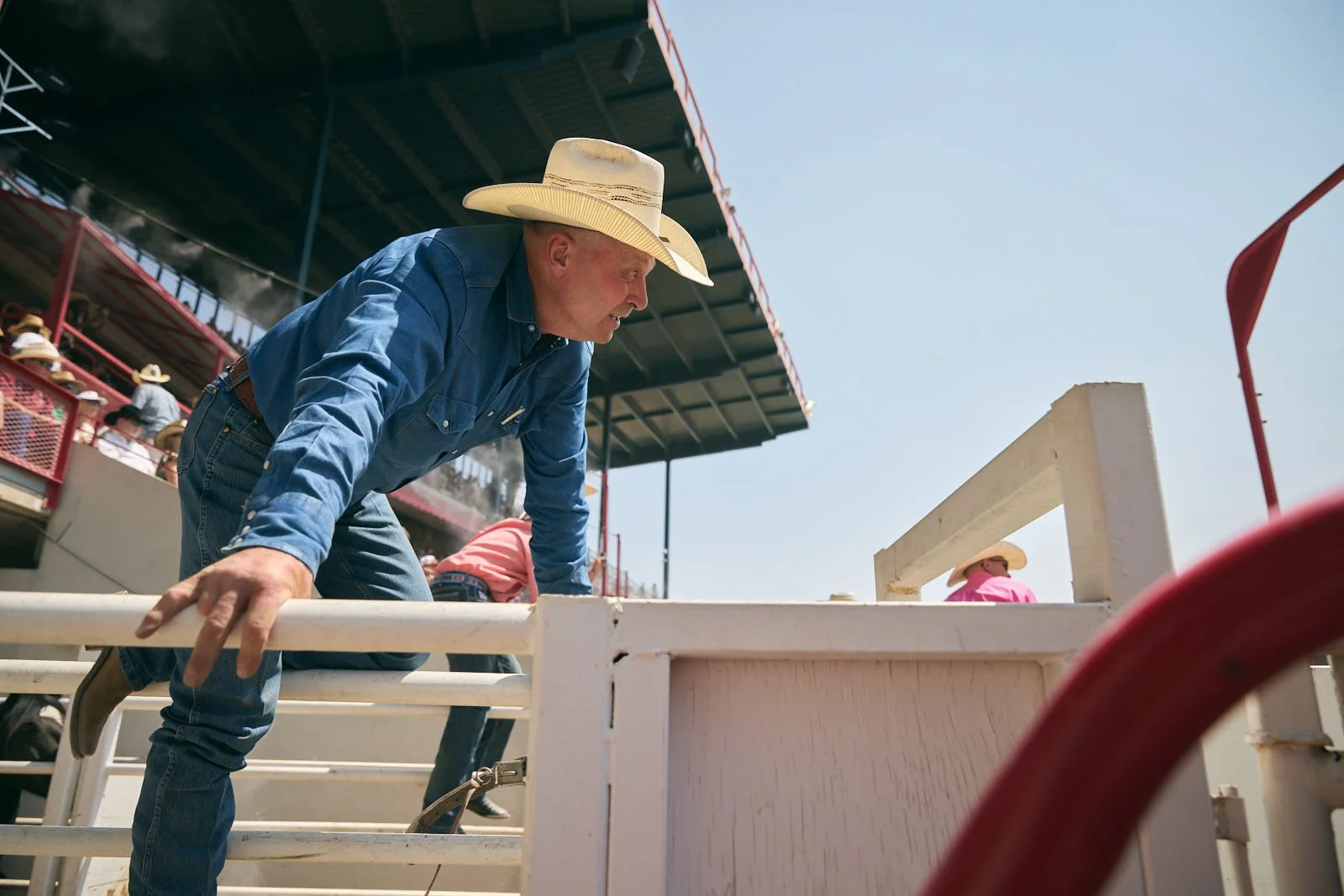 A man wearing a cowboy hat and denim shirt leaning over a fence at a rodeo arena with other spectators in the background.