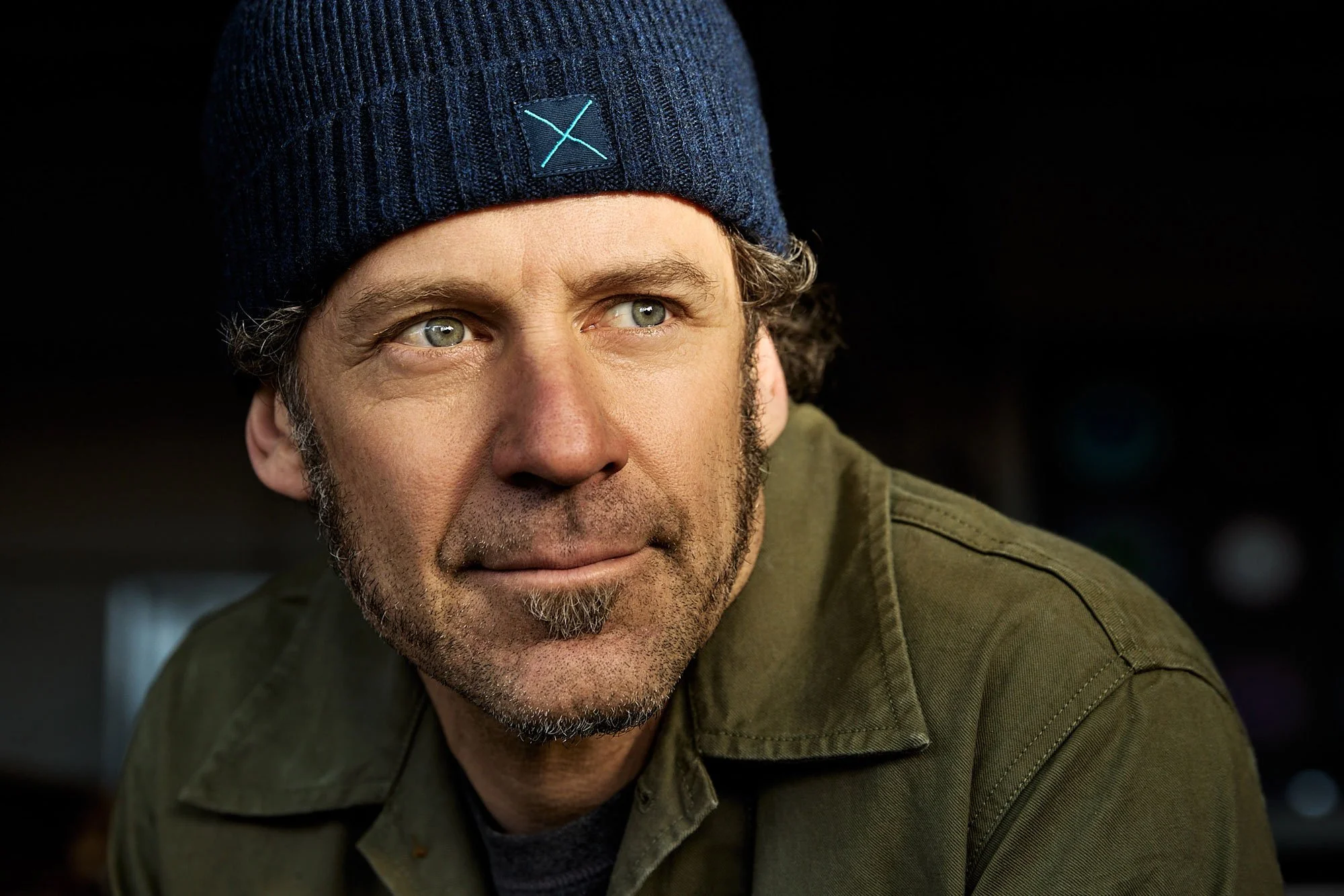 Close-up image of a man with light skin, short hair, facial hair, wearing a navy beanie and a green jacket, looking to the left.