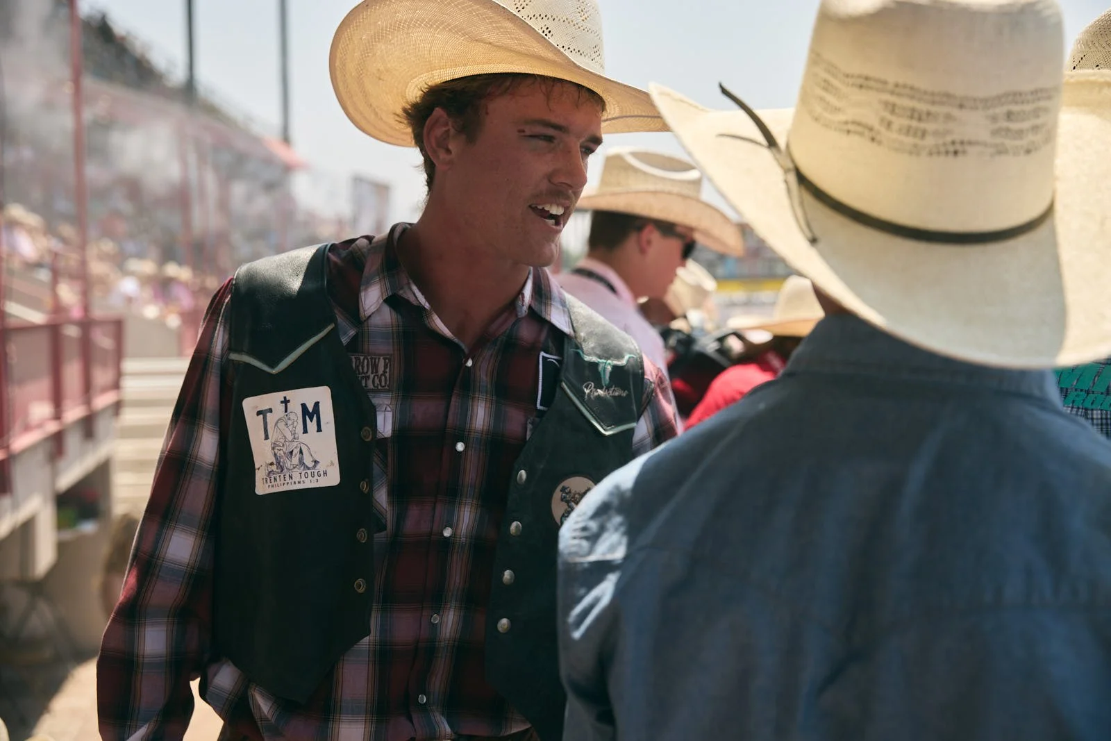 Two men wearing cowboy hats and plaid shirts talking outdoors, with others in the background at a fair or rodeo.