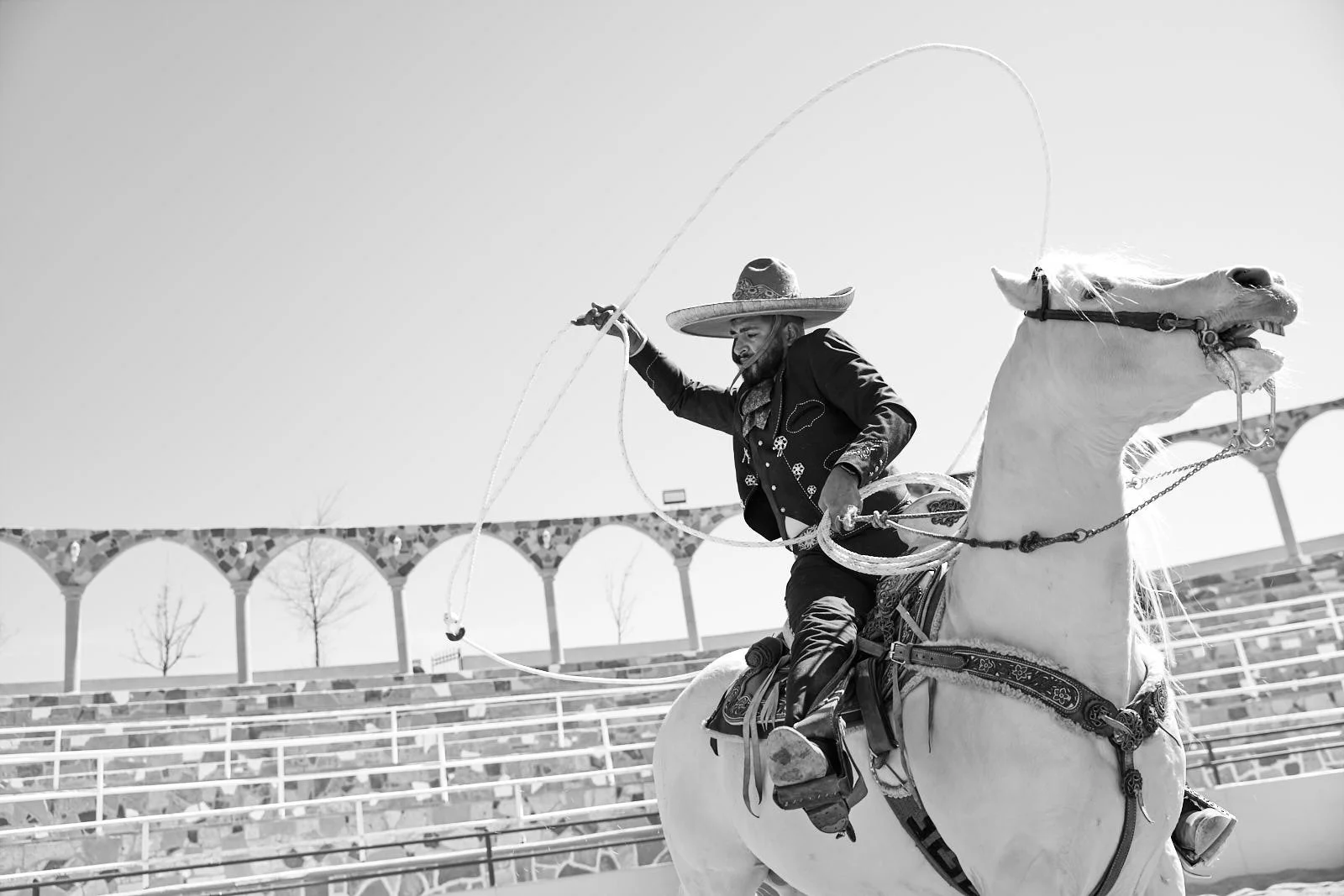 A man wearing a sombrero hat riding a white horse outdoors, holding a lasso in the air, with an arched bridge or structure in the background in a black-and-white photo.