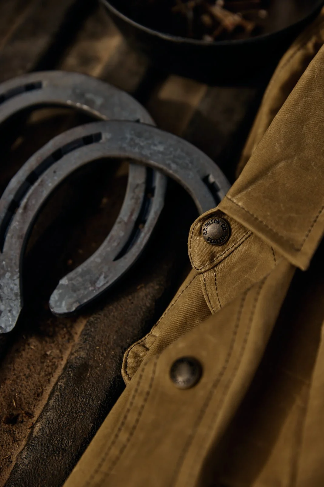 Close-up of metal horseshoes and a brown work jacket with a visible button. The horseshoes are placed on a wooden surface.