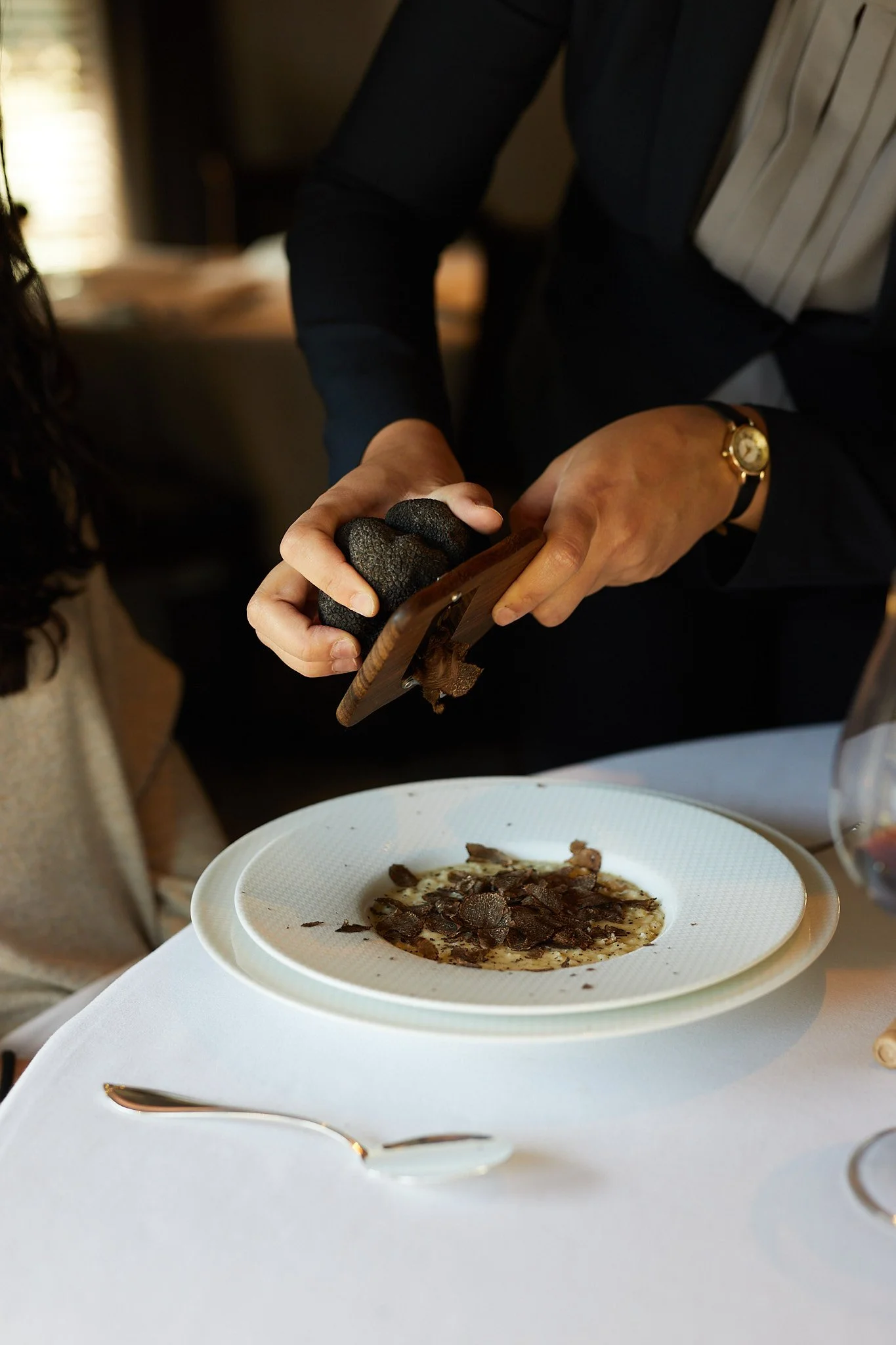 Person using a charcuterie knife to shave black truffle over a plate of sliced truffles on a white tablecloth.