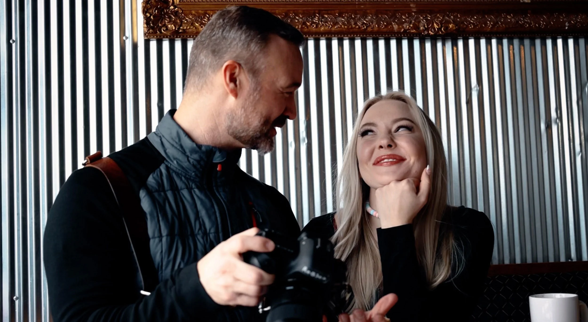 John Robson reviewing images with a woman inside a cafe with metallic wall and decorative wood trim.