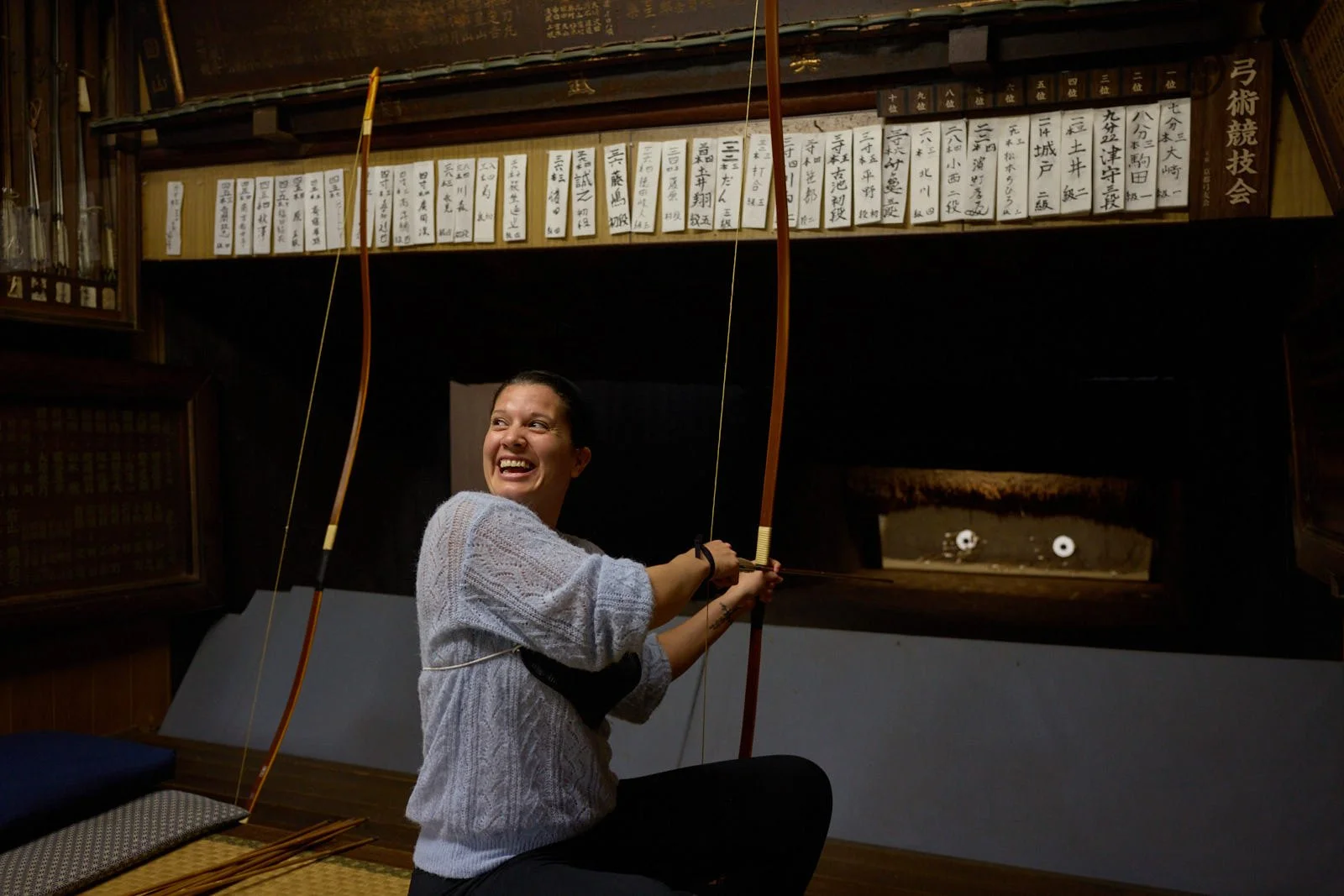 A woman smiling and aiming a traditional Japanese bow and wood bow indoors with wooden shelves and Japanese writing in the background.