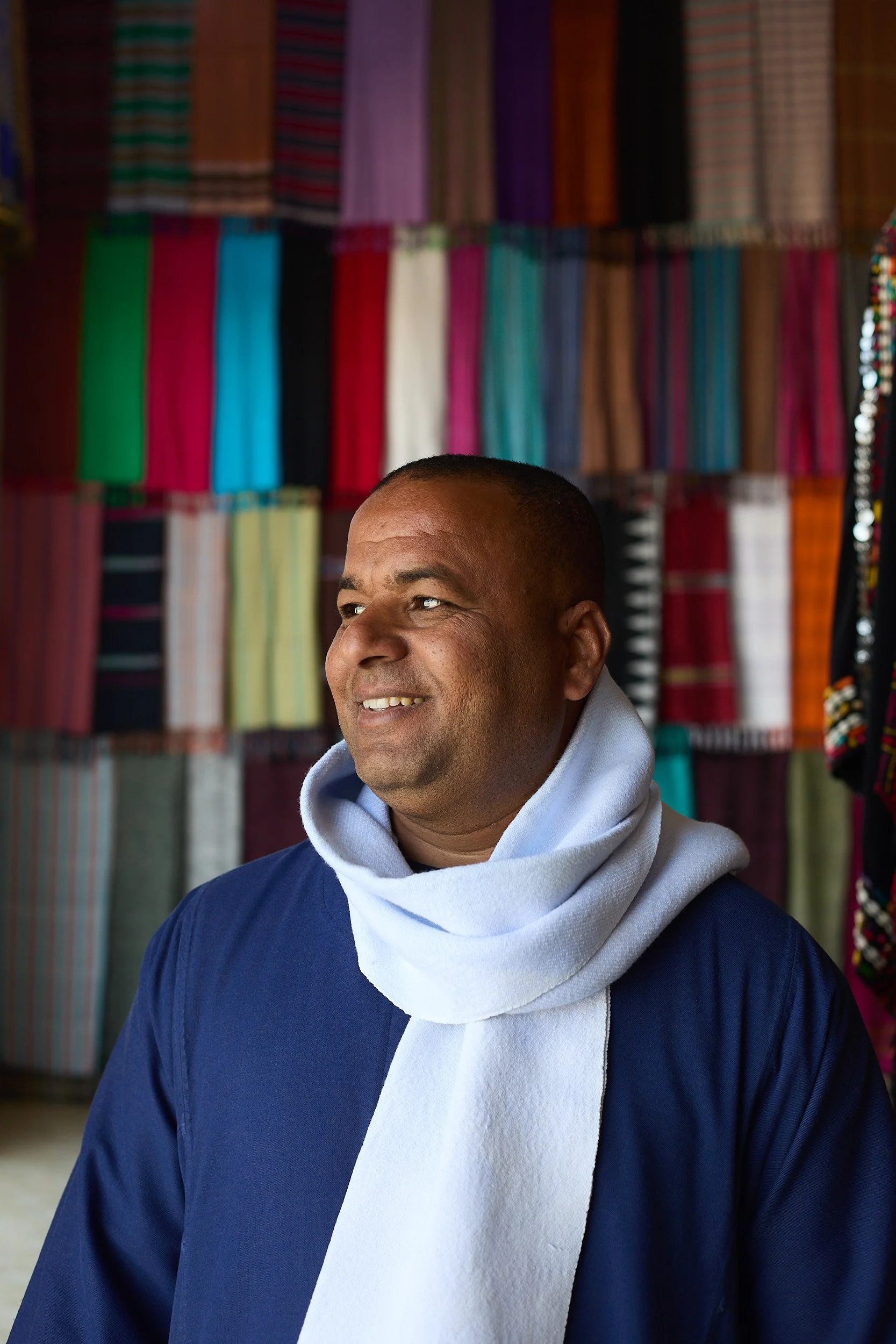 A man smiling while standing in front of a colorful fabric display, wearing a navy jacket and a light-colored scarf.