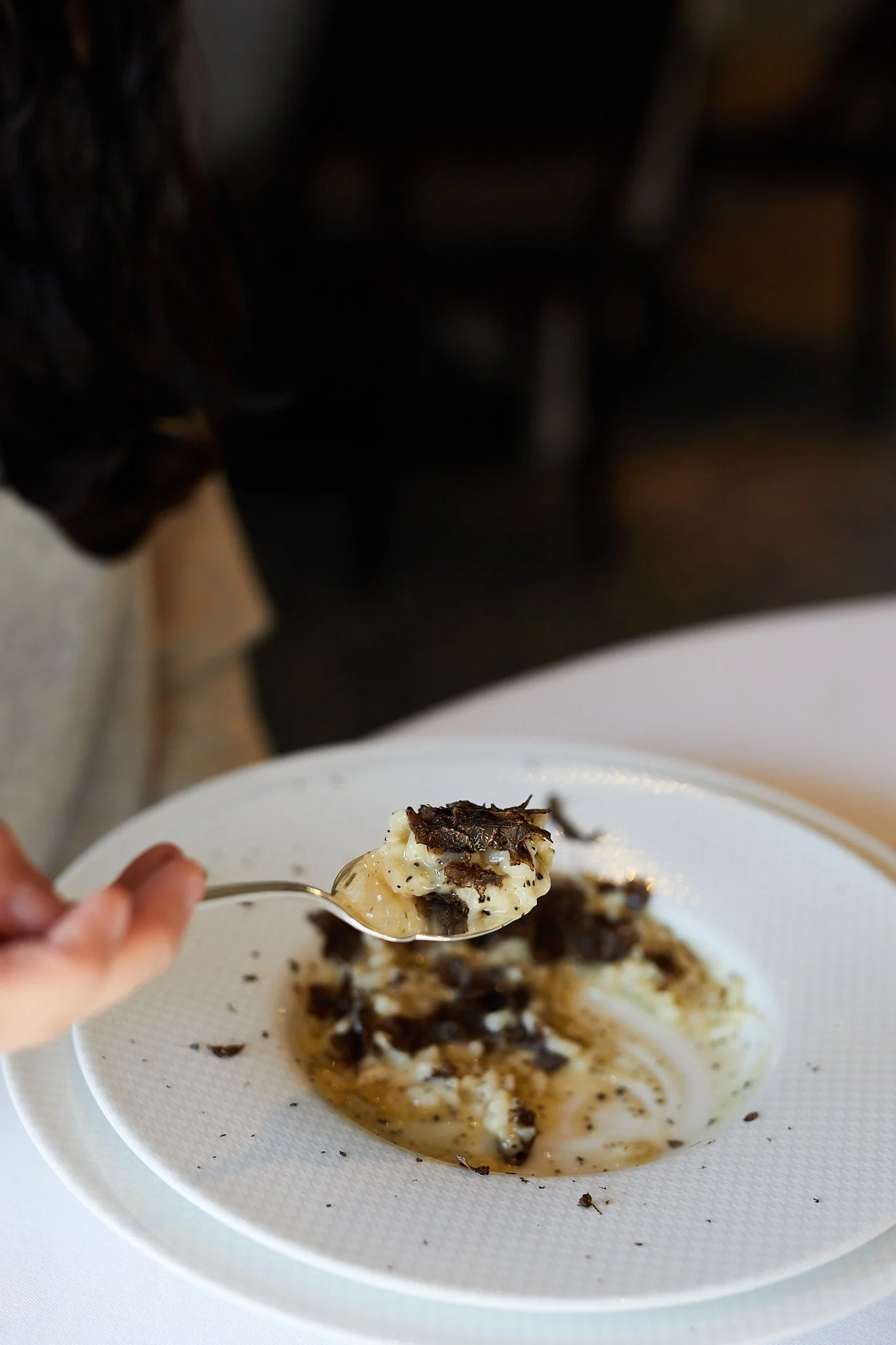 Close-up of a spoonful of creamy dish with black truffles, held over a white plate with more black truffles and sauce.