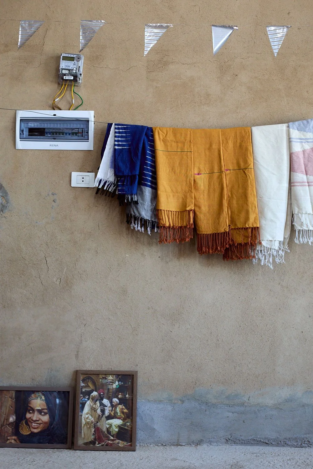 A beige wall with a string of colorful scarves hanging, a framed photograph of a woman in traditional attire, and a decorative metallic pennant banner