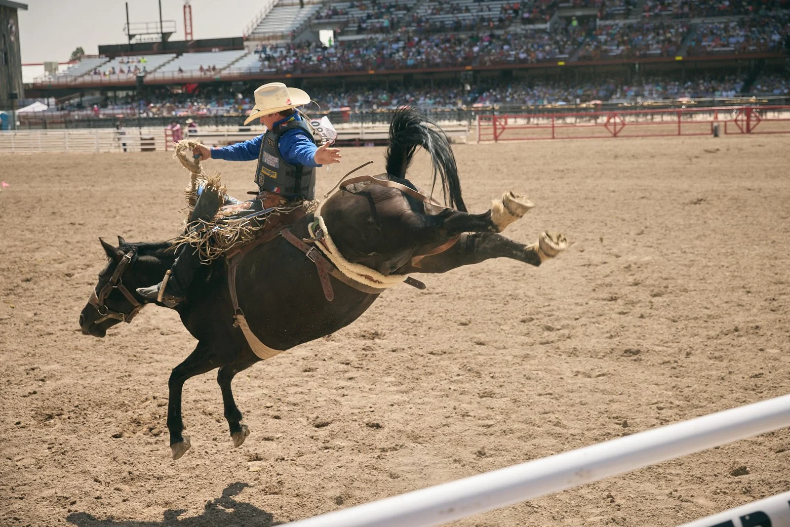 A rodeo cowboy riding a bucking horse in an arena, with a large crowd watching from the stands.