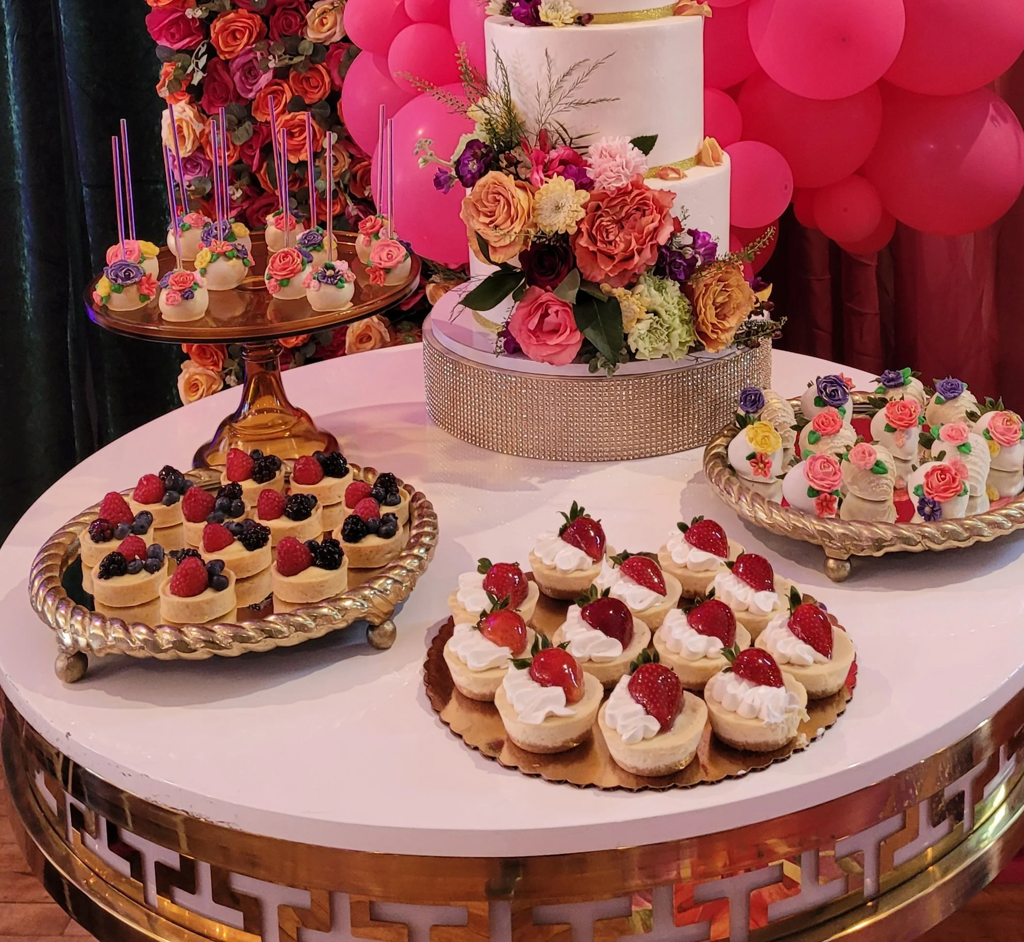 Three-tier white celebration cake decorated with fresh flowers, surrounded by trays of fruit tarts, strawberry cheesecakes, and floral cake pops.