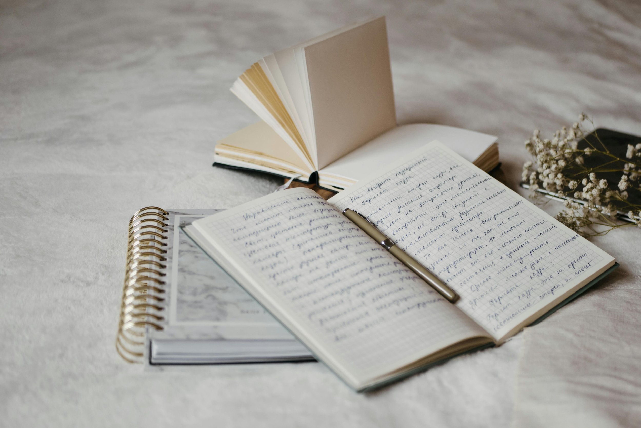 Open notebook with handwritten notes, a silver pen resting on it, a closed book with a black cover and floral sprigs beside it on a light-colored surface.