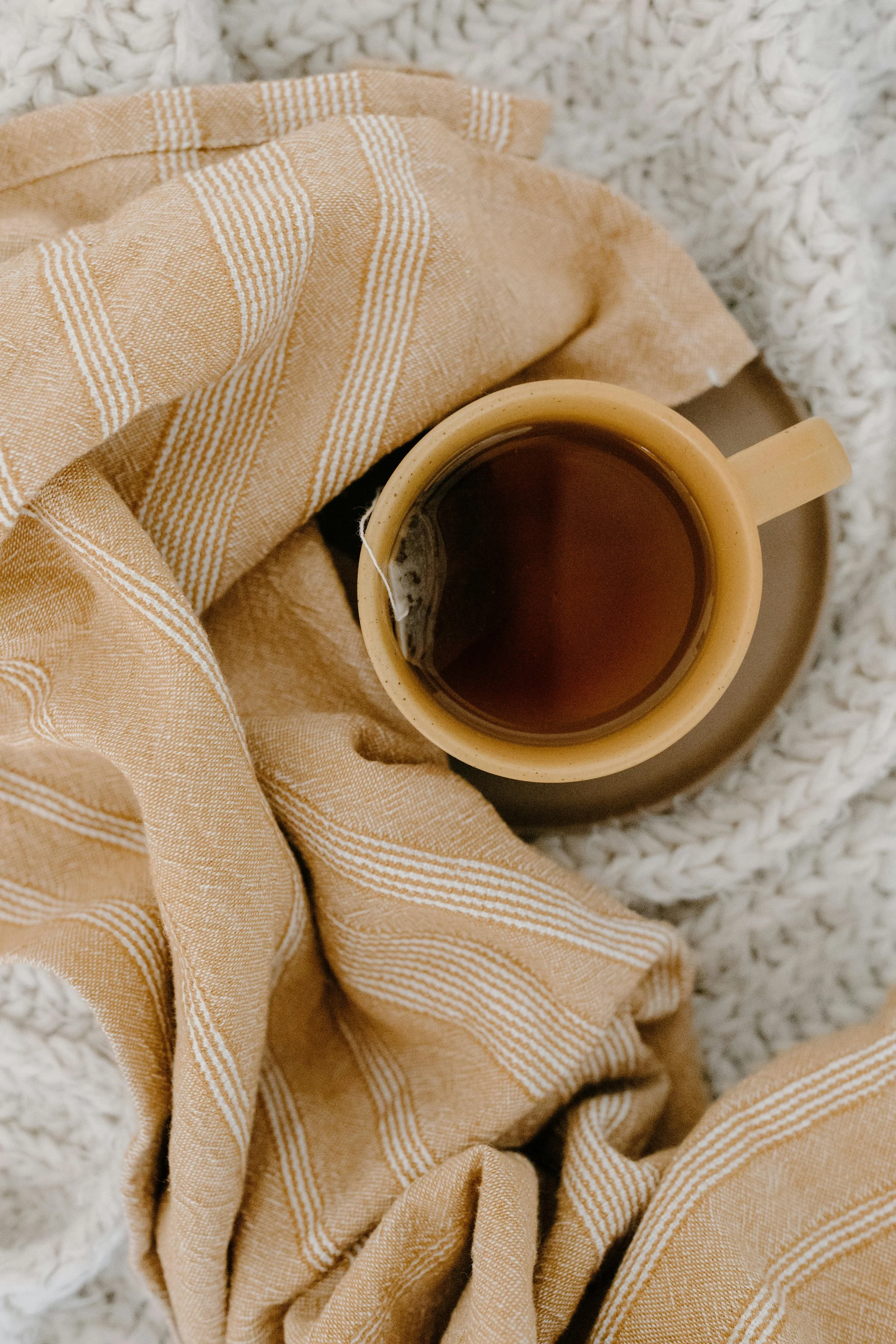 A beige and white striped blanket with a cup of black tea resting on it, on a textured white surface.