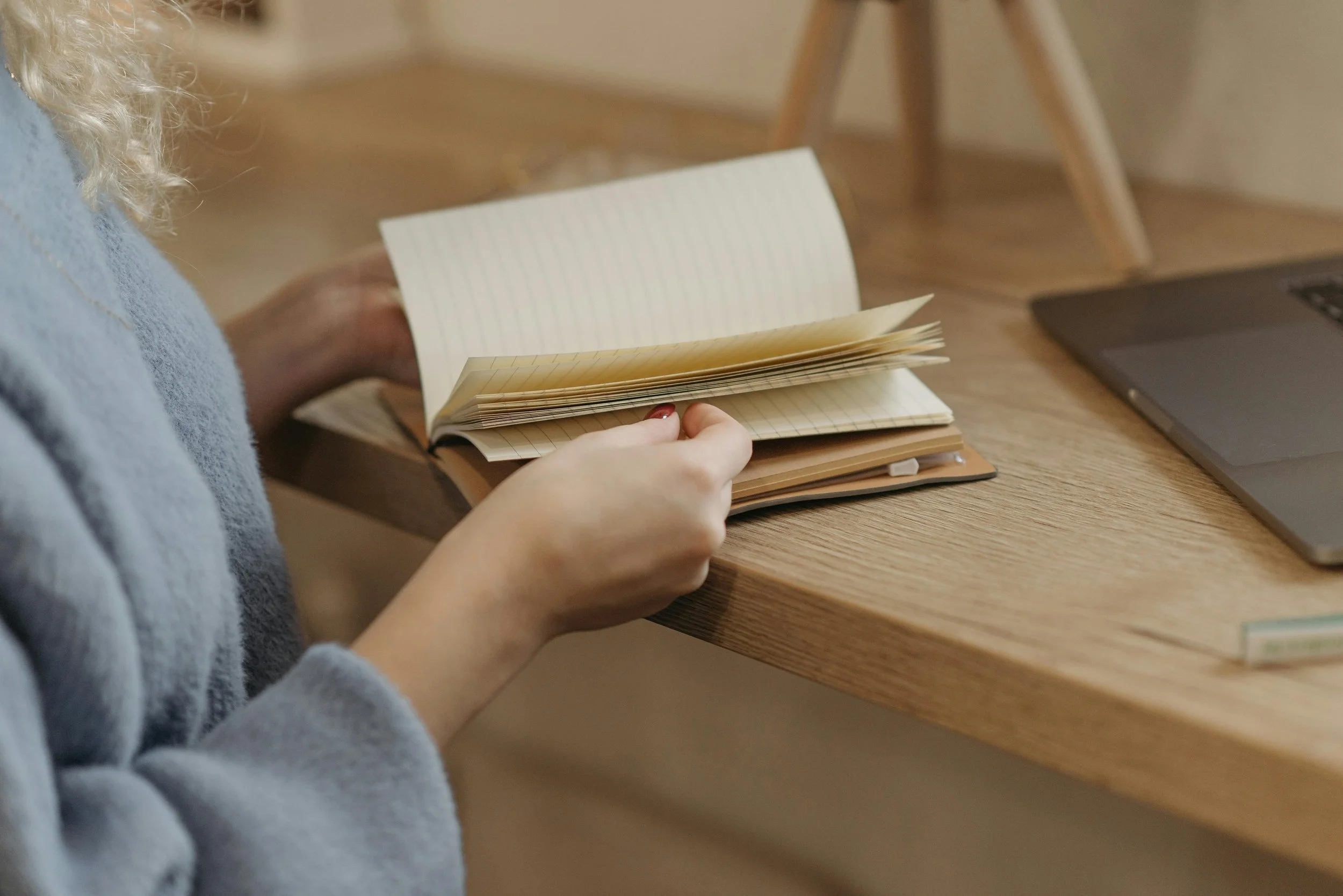 A person with curly blonde hair holding an open notebook with lined pages on a wooden table. A laptop is visible on the right side of the table.