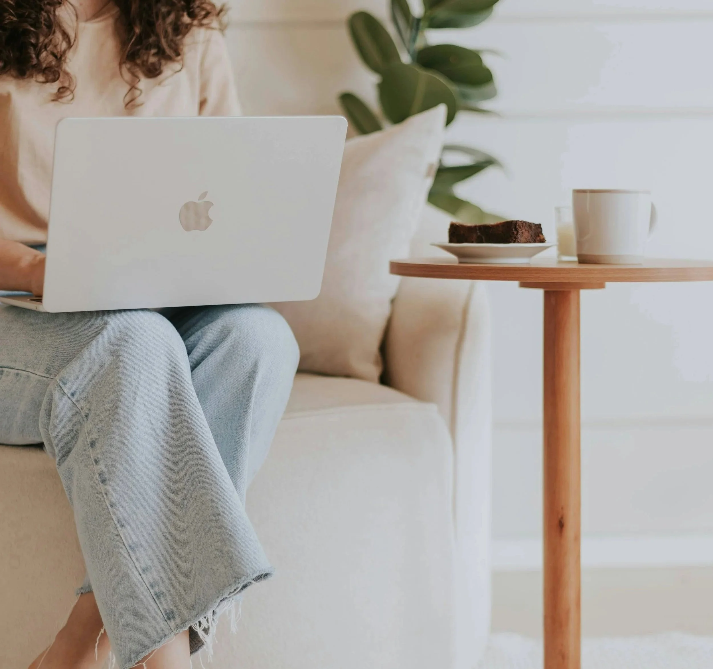 A woman sitting on a beige couch with curly brown hair, wearing light blue jeans with lace trim, holding a silver MacBook laptop. In front of her is a small wooden table with a chocolate cake on a white plate, a white mug, and a glass of milk. There is a large green leafy plant in the background near a window.