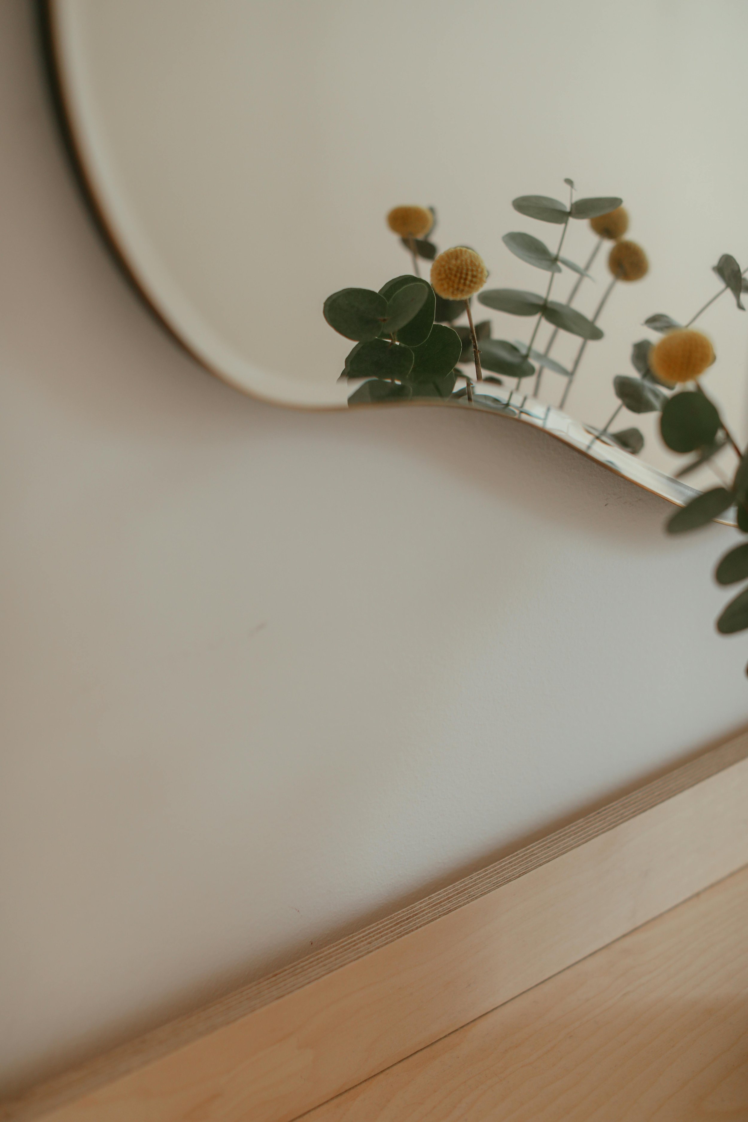 A close-up of a mirror reflecting a small bouquet of eucalyptus and yellow billy buttons flowers against a plain wall.