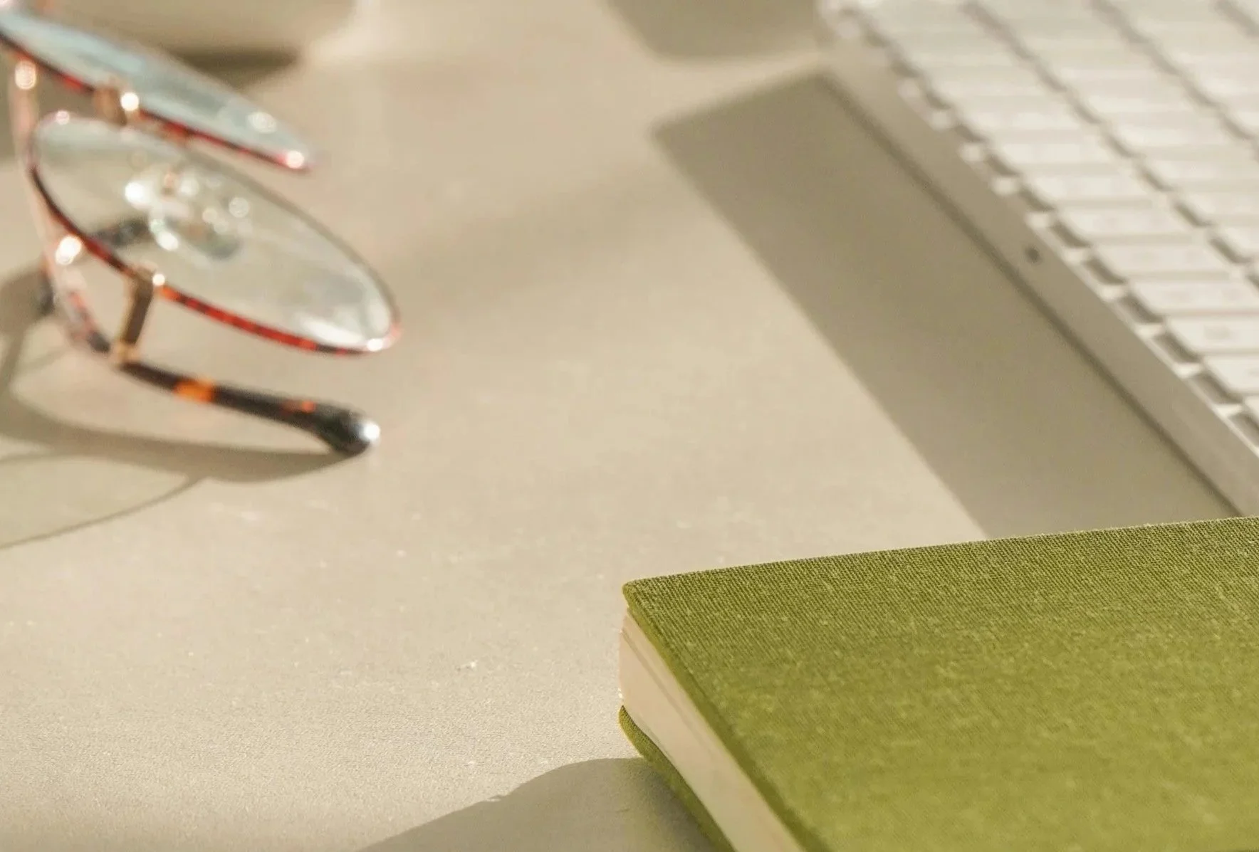 Close-up of a pair of eyeglasses with tortoiseshell frames on a beige desk, a green closed notebook, part of a silver laptop, and a green book in the foreground.