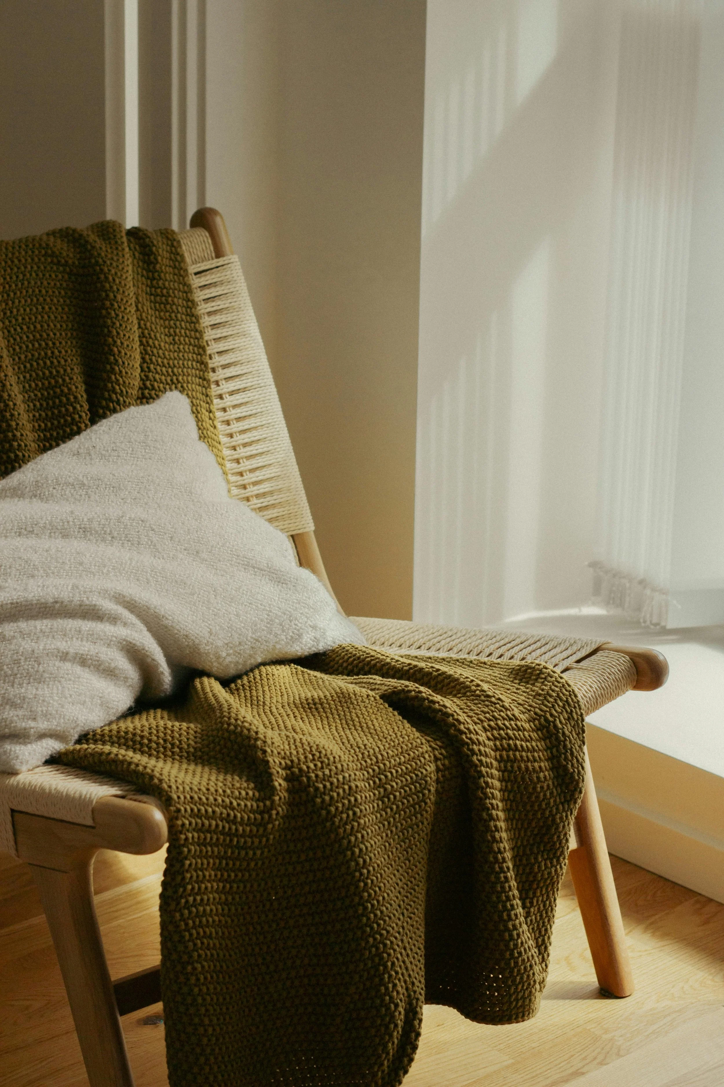 A wooden chair with a woven green blanket draped over it, a white textured pillow, and a mustard-colored textured blanket on the seat, positioned near a window with vertical blinds.