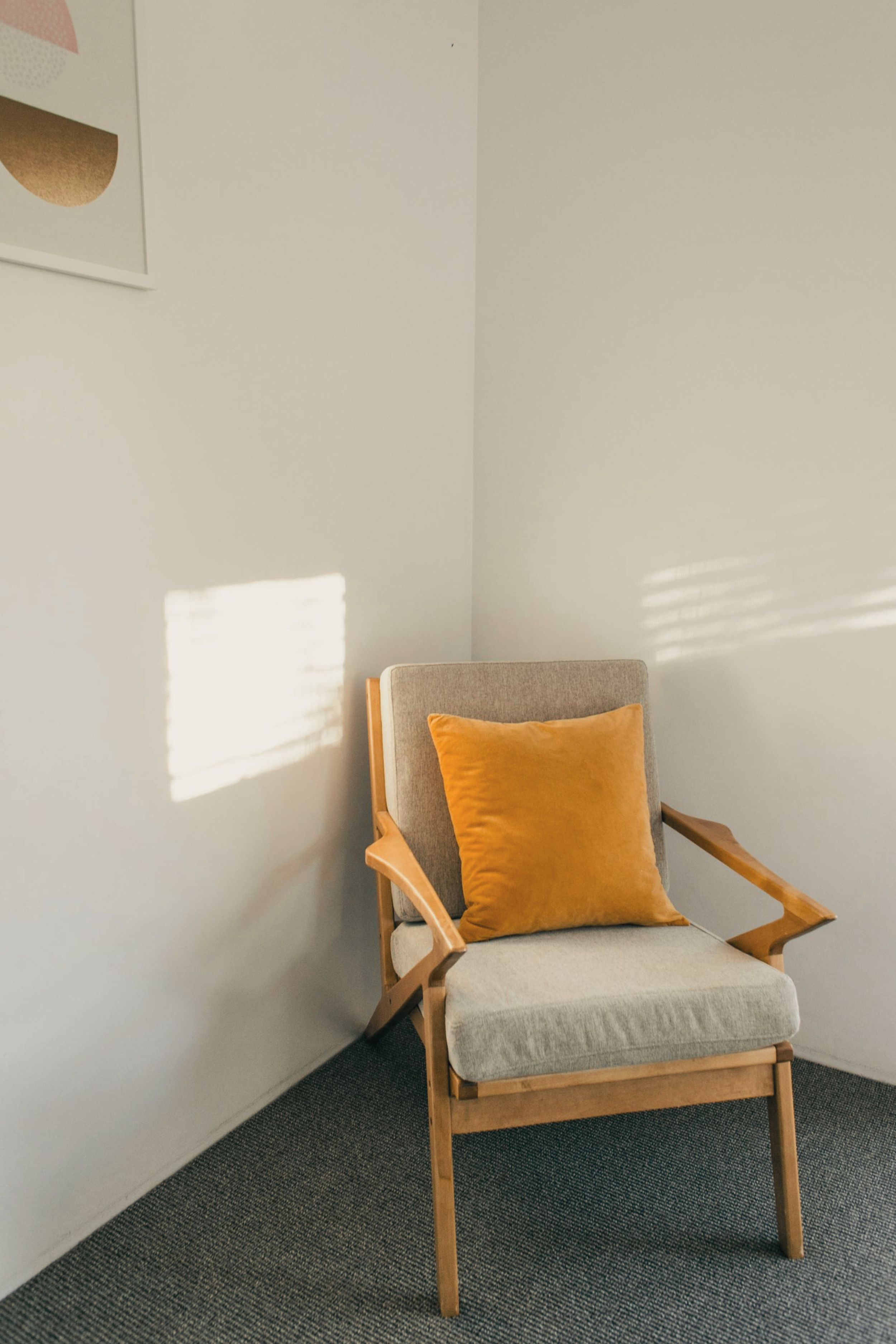 A modern armchair with light wood frame and gray fabric cushions, including a yellow-orange velvet pillow, situated next to a plain off-white wall with sunlight creating a pattern of light and shadow on it.
